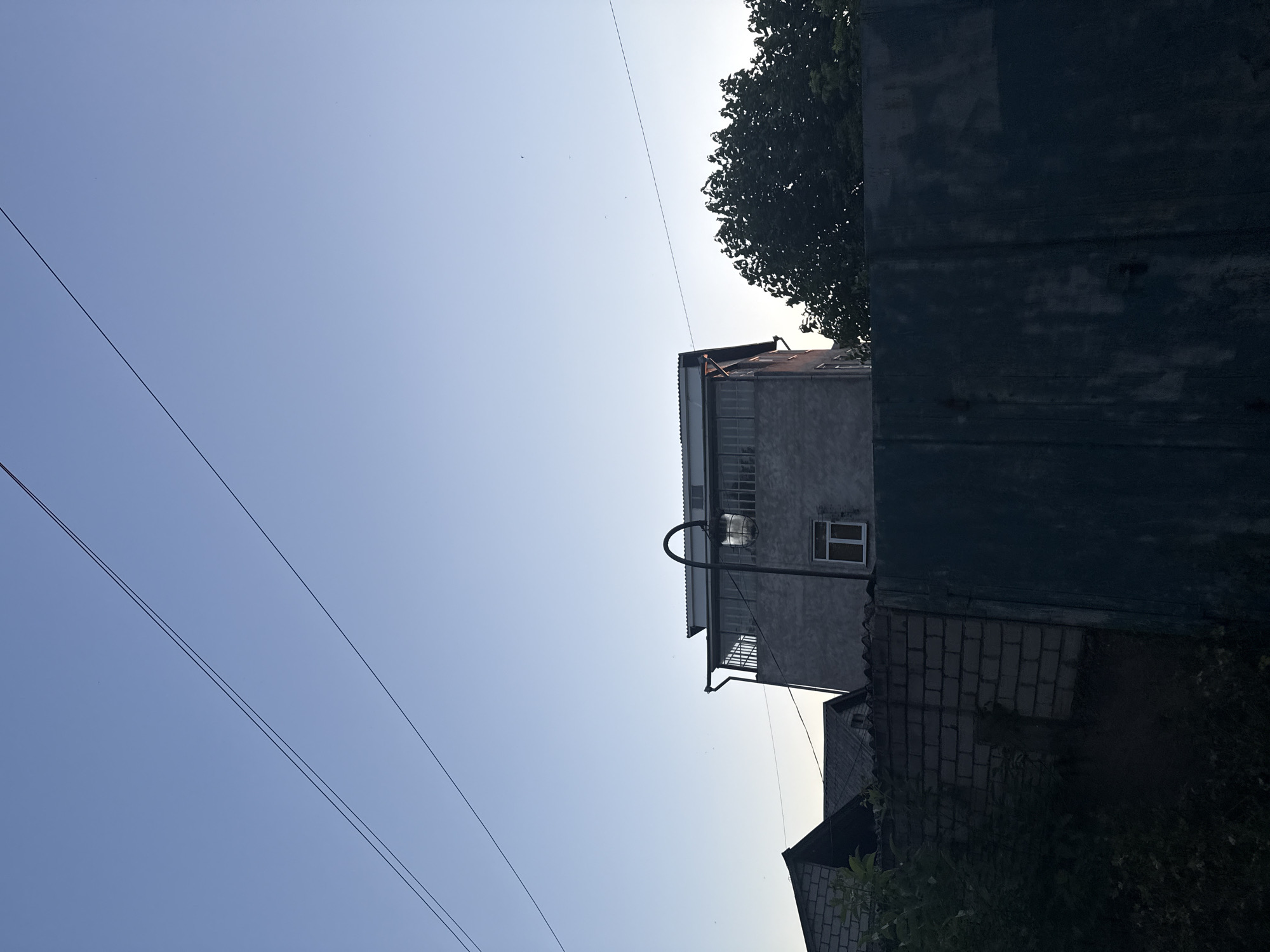 The image depicts a residential area in Obukhivka, Ukraine, under a clear blue sky during twilight. In the foreground, a dark, weathered fence dominates the lower portion of the frame. Behind the fence, a low brick wall is visible, partially obscured by dense green foliage. Further back, several houses are present. To the left, a brick house with a dark roof stands partially illuminated. In the center, a two-story building with a concrete facade features a balcony with a metal railing and a large window. A street lamp with a globe-shaped light fixture is mounted on a curved pole in front of this building. To the right, the dark canopy of a lush tree rises above the fence line. Electrical wires stretch across the sky, crisscrossing the scene. The overall atmosphere is serene, suggesting a quiet evening in a rural or suburban setting.