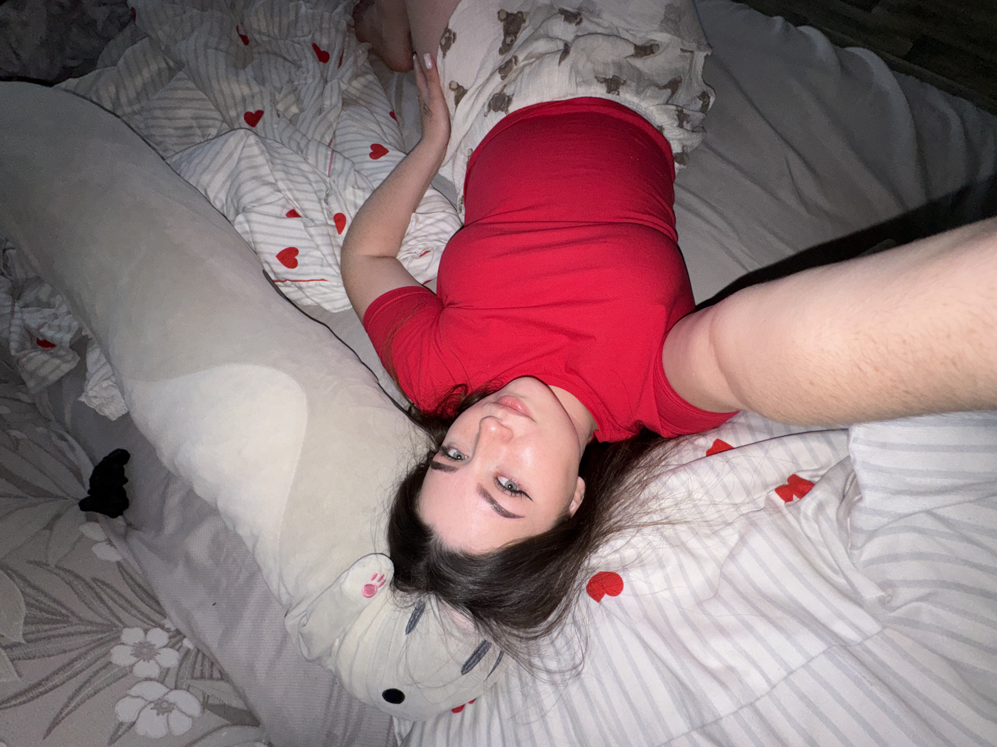 An overhead, close-up shot captures a young woman lying on her back on a bed, taking a selfie. She is positioned towards the left side of the frame, looking directly upwards at the camera with a neutral expression. Her dark hair is splayed around her head on the pillow, and she wears prominent winged eyeliner and light makeup. She is dressed in a plain red t-shirt and light-colored pajamas or shorts patterned with small brown bear figures.
Her left arm is bent at the elbow, with her hand resting near her head, while her right arm is extended upwards, presumably holding the device used to take the photo. A large, light grey plush body pillow, resembling a rabbit or bear with stitched features and a pink paw print on one ear, lies alongside her on the bed, extending from near her head down to her legs.
The bedding consists of a duvet cover with light grey horizontal stripes and scattered red heart motifs. The bottom sheet or fitted sheet visible beneath has a pattern of white flowers on a light grey background. The lighting is dim, suggesting an indoor setting during evening or nighttime. The overall scene conveys a relaxed, personal moment in what appears to be a bedroom in Shirkovo, Russia. There is no visible text.