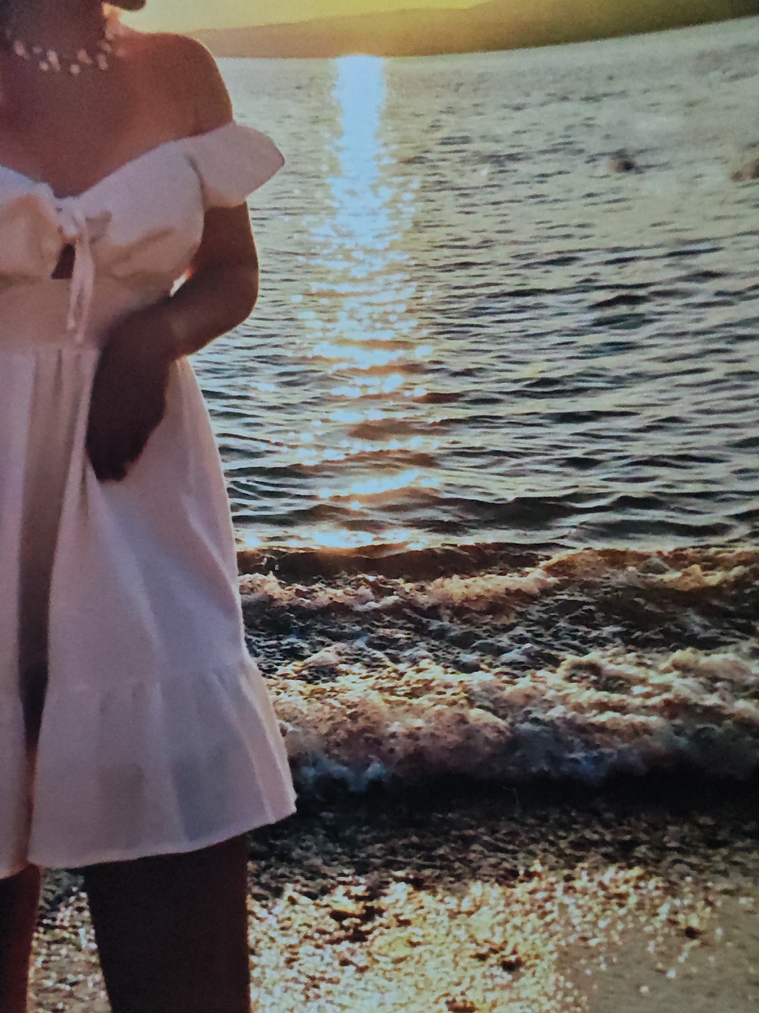 The image shows the lower half of a person wearing a white off-the-shoulder dress standing on a sandy beach. The person is wearing a pearl necklace. The ocean is in the background with the sun setting, creating a reflection on the water. Waves are breaking on the shore.