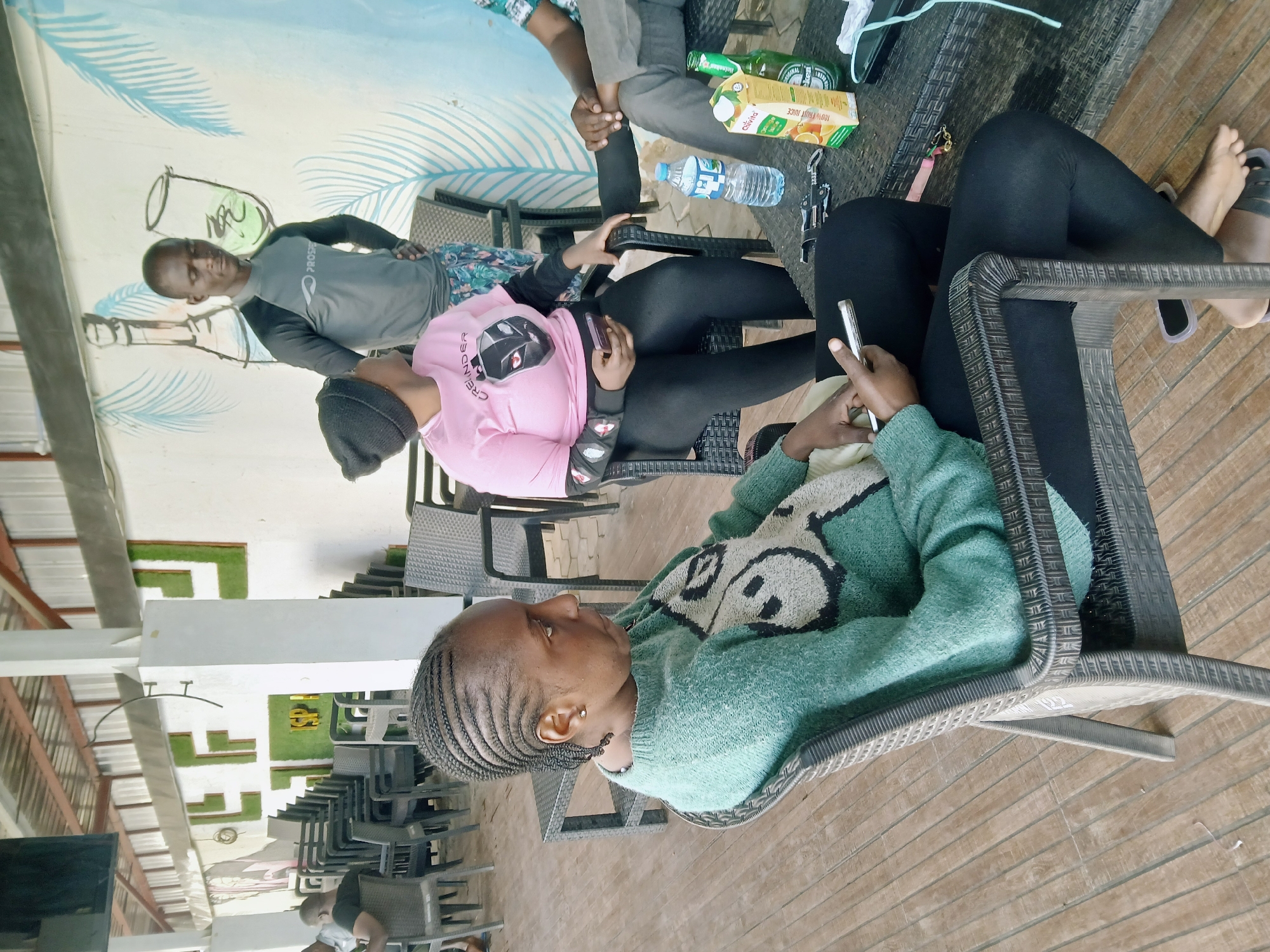 A medium shot captures a group of four individuals in an outdoor setting in Barakin Lerawi, Nigeria.
One individual, wearing a light green sweater, is seated in a gray wicker chair. This person has braided hair and is holding a silver smartphone.
Another individual wears a pink long-sleeved shirt, black leggings, and a black beanie. This person holds a black smartphone.
A third person wears a gray rash guard and sits with one hand on a table.
A fourth person wears light-colored pants and a buttoned shirt.
A dark gray wicker table holds a green beer bottle, an orange juice box, and a water bottle.
The background shows a wall painted with tropical-themed images, including blue palm leaves. The setting is in an open-air covered patio.
