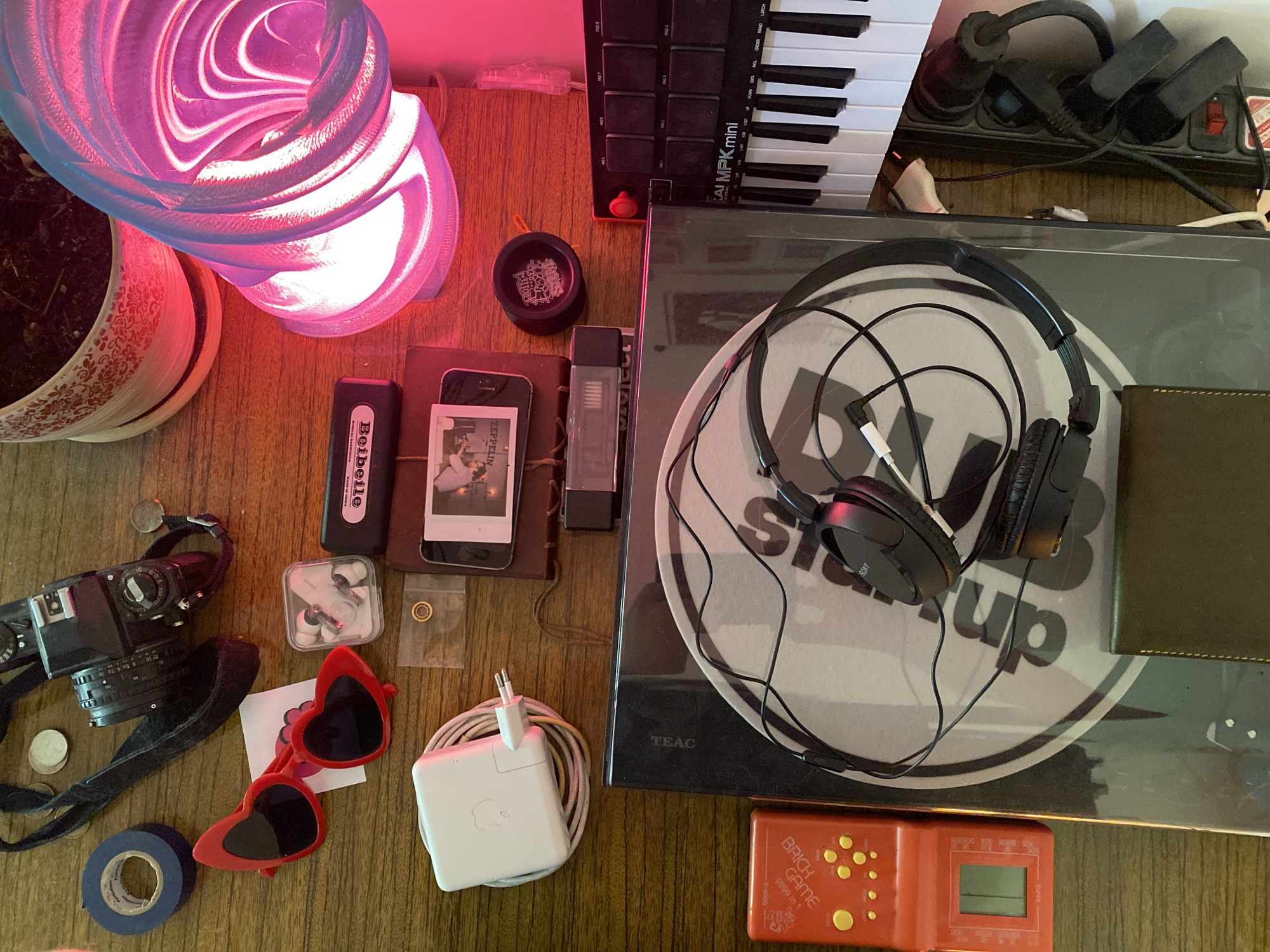 This is a cluttered tabletop scene. Starting from the left, there's a potted plant, a modern lamp emitting a pinkish-red light, a film camera with a black strap, some coins, blue electrical tape, red heart-shaped sunglasses, a pair of wireless earbuds in a case, a small bag with a bearing, a phone in a brown case with a Polaroid photo tucked into it, a battery pack, a black yo-yo, a small gray storage drive, an Apple laptop power adapter with coiled cable, a black AKAI MPK mini MIDI keyboard, a power strip, black Sony headphones, a green wallet or notebook, a TEAC record player, and an orange handheld "Brick Game" console.
The location in Rusanovo, Russia, cannot be determined.