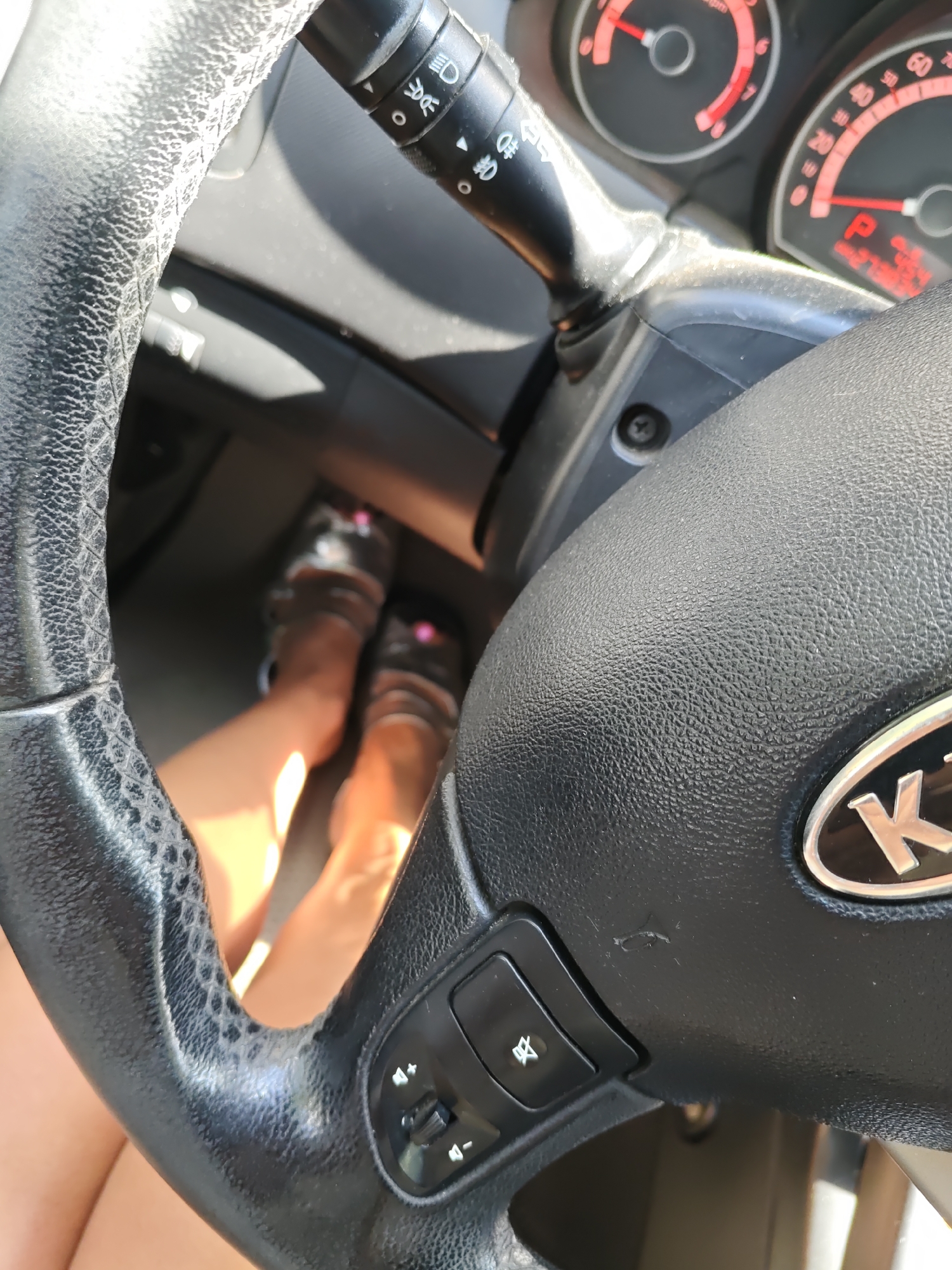 A first-person perspective captures the interior of a car, focusing on the steering wheel and the driver's feet. The steering wheel is black and textured, with integrated controls for volume and cruise control on the right side. A stalk for windshield wipers and turn signals is visible to the left of the steering column. The dashboard features a red-lit speedometer and other gauges, one of which indicates "P" for park. The driver's legs and feet, clad in sandals, are visible in the lower portion of the image, resting on the pedals. The overall lighting suggests a sunny day. The specific location in Rostov-on-Don, Russia, cannot be determined from the visual information alone.