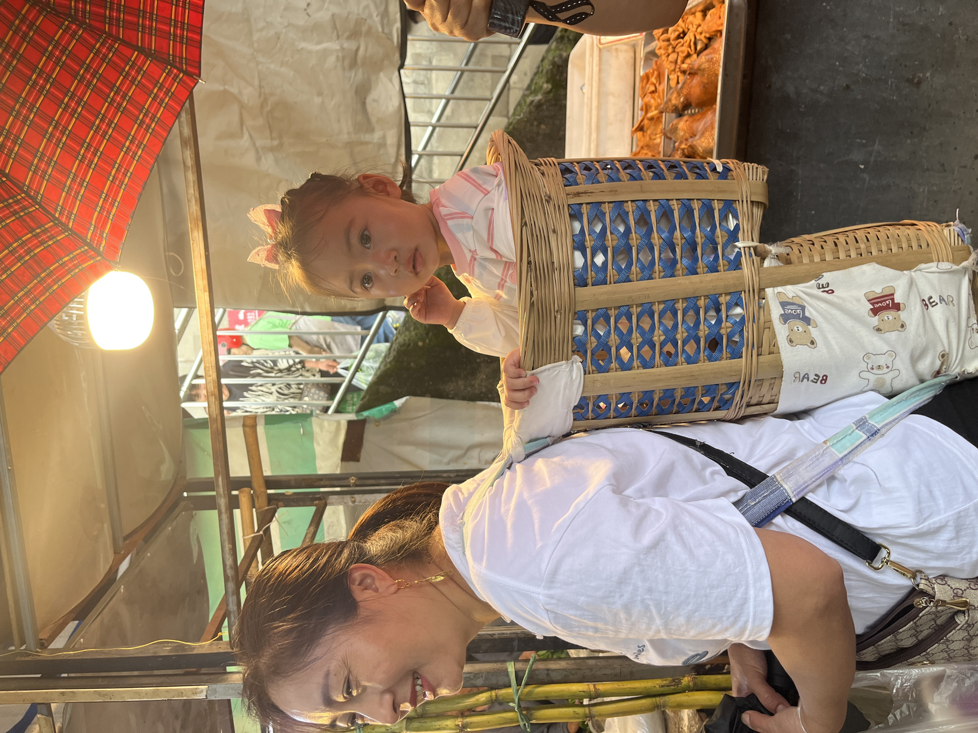 This image captures a candid scene in an outdoor or semi-enclosed market in Jiangjin, China. The primary subjects are an adult woman carrying a young child on her back.
The woman, positioned in the left foreground and central, is seen from the mid-chest up in a three-quarter profile facing left. She has dark hair pulled back in a ponytail and wears a white short-sleeved shirt. A pair of gold-toned, leaf-shaped earrings are visible. Her mouth is open in a smile, and her gaze is directed beyond the left frame. A dark strap from a shoulder bag or the child-carrying apparatus crosses her right shoulder. A beige shoulder bag with a brown patterned design (resembling a "Gucci" monogram) is visible on her right hip.
A young girl, estimated to be between one and three years old, is seated upright in a woven bamboo basket strapped to the woman's back. The child is positioned to the right of the woman's head and shoulders. She has dark hair gathered on top of her head with a pink bow headband and wears a long-sleeved white shirt with light pink stripes. Her expression is neutral, and her gaze is directed towards the right-middle distance. Her right hand is raised near her cheek. The basket itself is constructed from natural bamboo, with a prominent blue woven pattern forming diamond shapes on its exterior. A white fabric, patterned with cartoon bears and text reading "LOVE BEAR," is tucked into the lower front interior of the basket.
The background reveals elements of a bustling market environment. Overhead, a bright, bare light bulb hangs from a metal frame, providing illumination. A large, open umbrella with a red and black plaid pattern is partially visible in the upper right quadrant. Below the umbrella, a metal tray filled with various cooked food items, appearing golden-brown and possibly fried, is displayed on a counter. To the far left, several tall, green sugarcane stalks are bundled together. Beyond the subjects, blurred figures suggest other individuals, including one in a green shirt and white apron in the middle-left background. Metal railings and what appear to be steps are visible in the far background, indicating multi-level structures or pathways. The overall setting is characterized by temporary structures with metal framing and light-colored sheeting overhead.