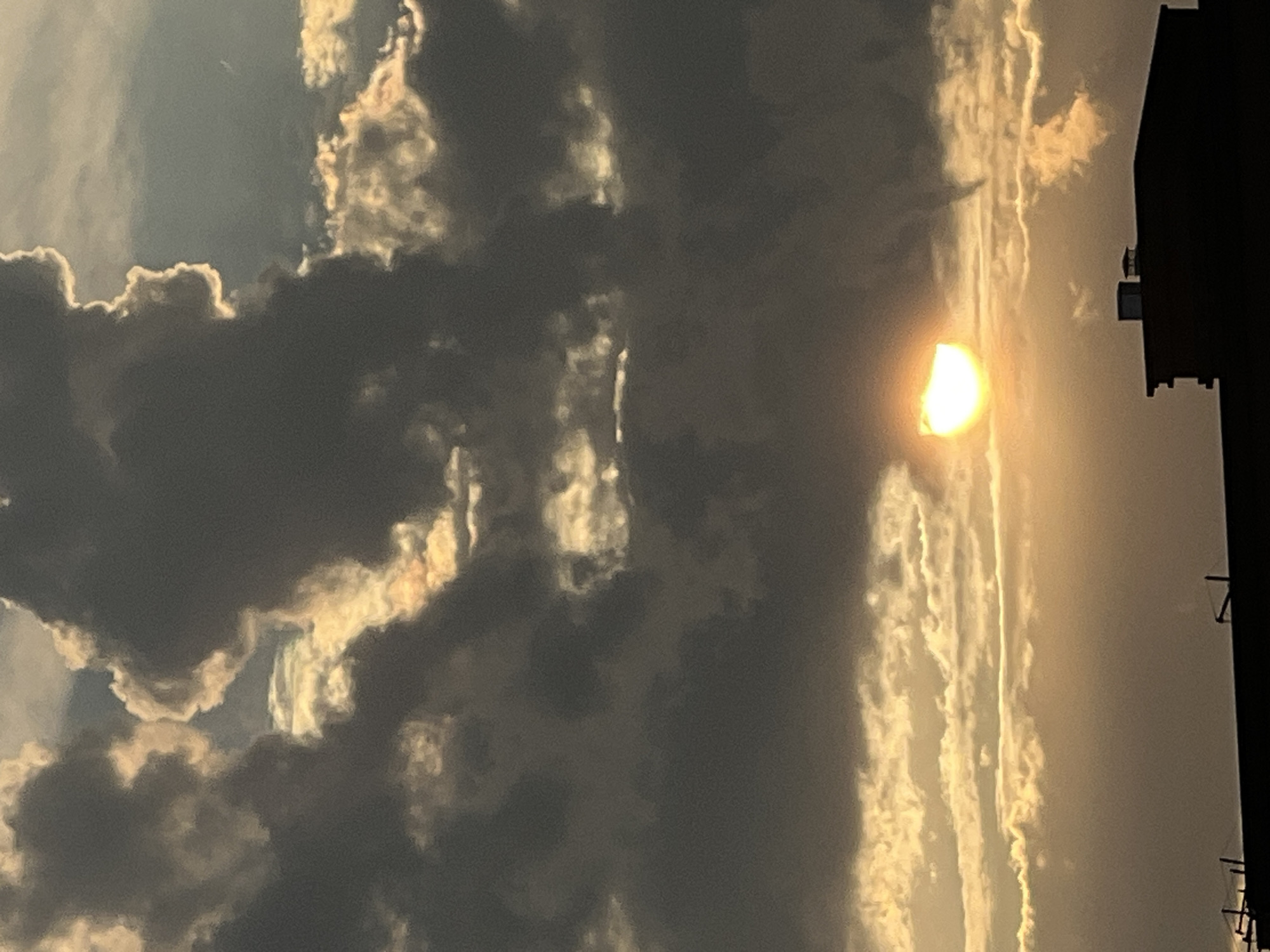 A photograph captures a dramatic sky scene with the sun partially obscured by dark, heavy clouds. The sun, a bright, semi-circular orb, casts a warm, golden glow through a break in the clouds, illuminating the edges of surrounding cloud formations with a luminous, almost ethereal light. These clouds appear textured and dense, with darker, shadowed areas contrasting sharply with the brightly lit portions. To the right of the frame, a dark, silhouetted building or structure is visible, suggesting an urban or suburban setting. The overall impression is of a late afternoon or early evening sky, possibly with an approaching storm or the aftermath of one, given the dramatic interplay of light and shadow. The location context of Krasnodar, Russia, provides a geographical anchor for this atmospheric display. There is no visible text in the image.