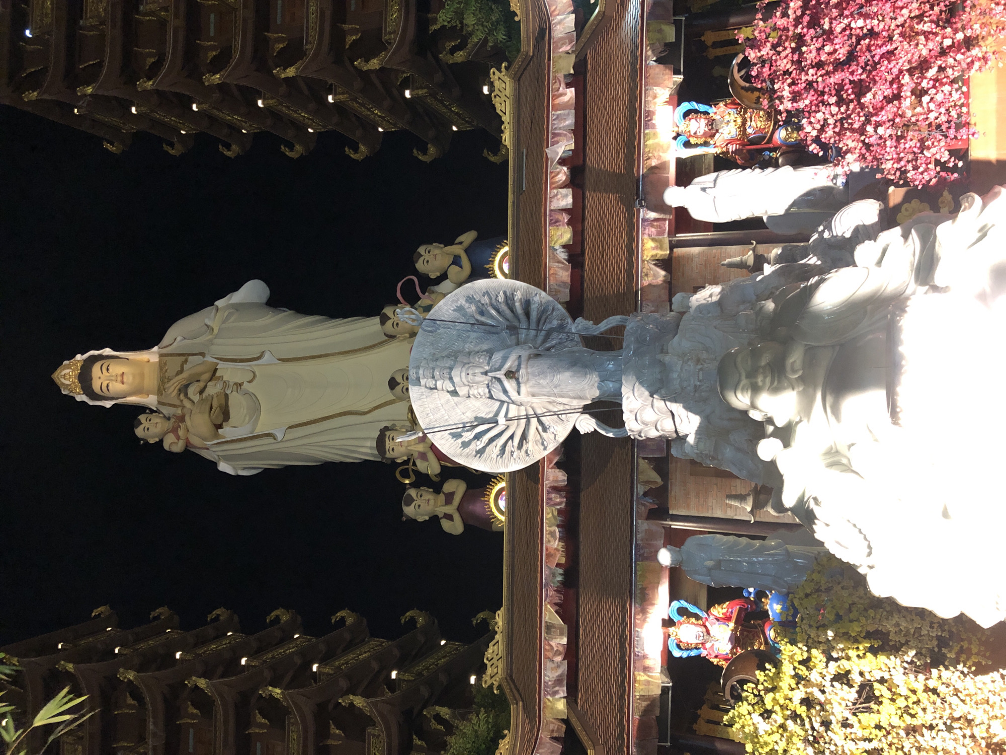 This image depicts the Linh Son Truong Tho Pagoda in Mui Ne, Vietnam, at night. The focal point is a large, illuminated white statue of a deity, possibly Guanyin, holding a child. Flanking the pagoda are two multi-tiered, pagoda-style buildings with intricate gold trim and illuminated windows. Below the central deity statue, there is a white, multi-armed stone sculpture. In front of this, a large, white, carved figure, possibly a Buddha, is partially visible and brightly lit. The foreground features vibrant displays of yellow and pink flowers, with a smaller, colorful statue of a warrior deity to the right. The scene is illuminated by artificial lighting, creating a dramatic contrast between the dark sky and the brightly lit structures and statues.