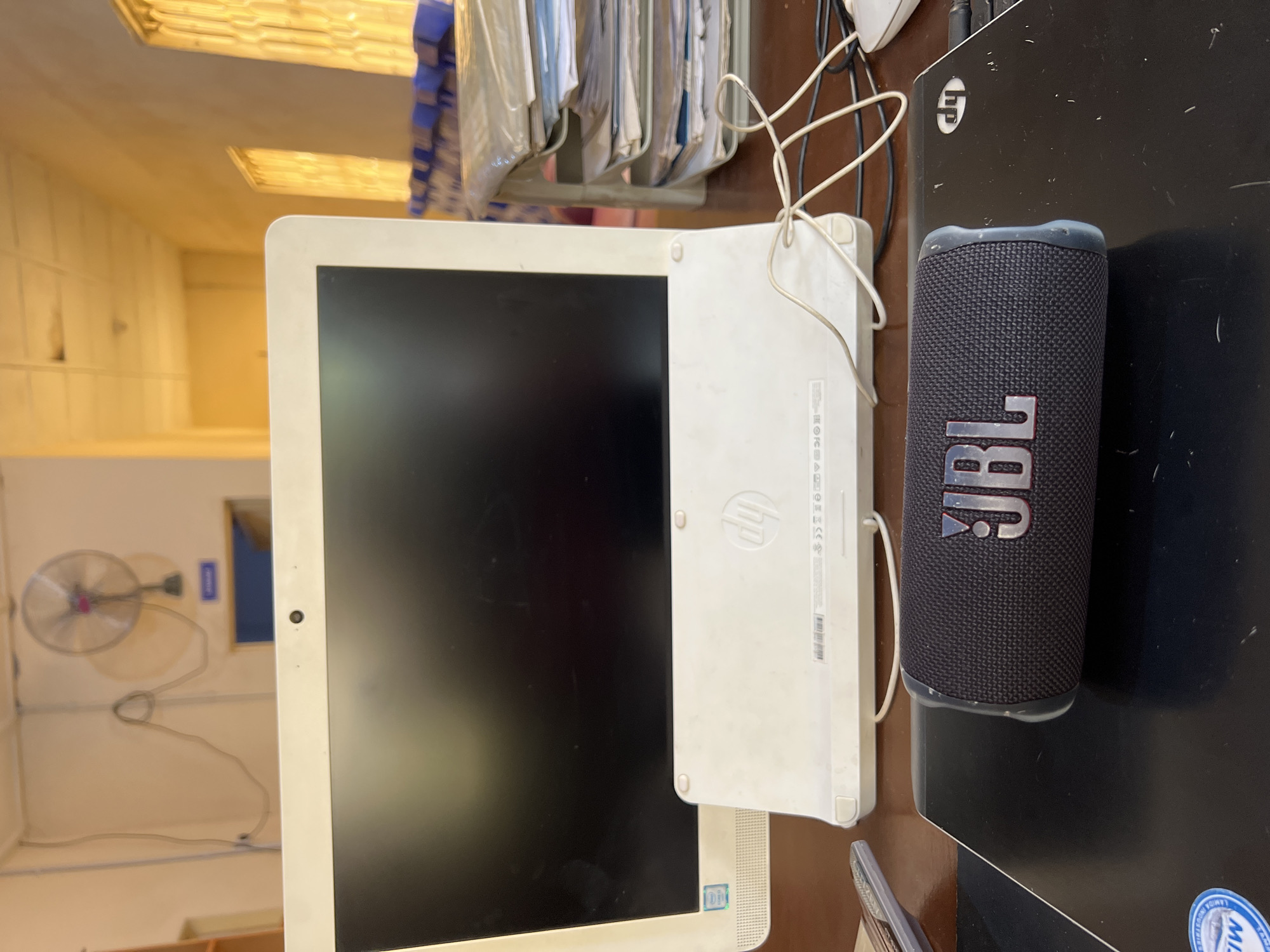 The image depicts a desktop computer, a JBL speaker, and a laptop on a desk. The desktop is white and has a black screen. The JBL speaker is black with the JBL logo in silver. The laptop is black. In the background is a wall-mounted fan in a room with light yellow walls. No people are visible. There is no verifiable information to confirm that this is in the city of Jos, Nigeria.