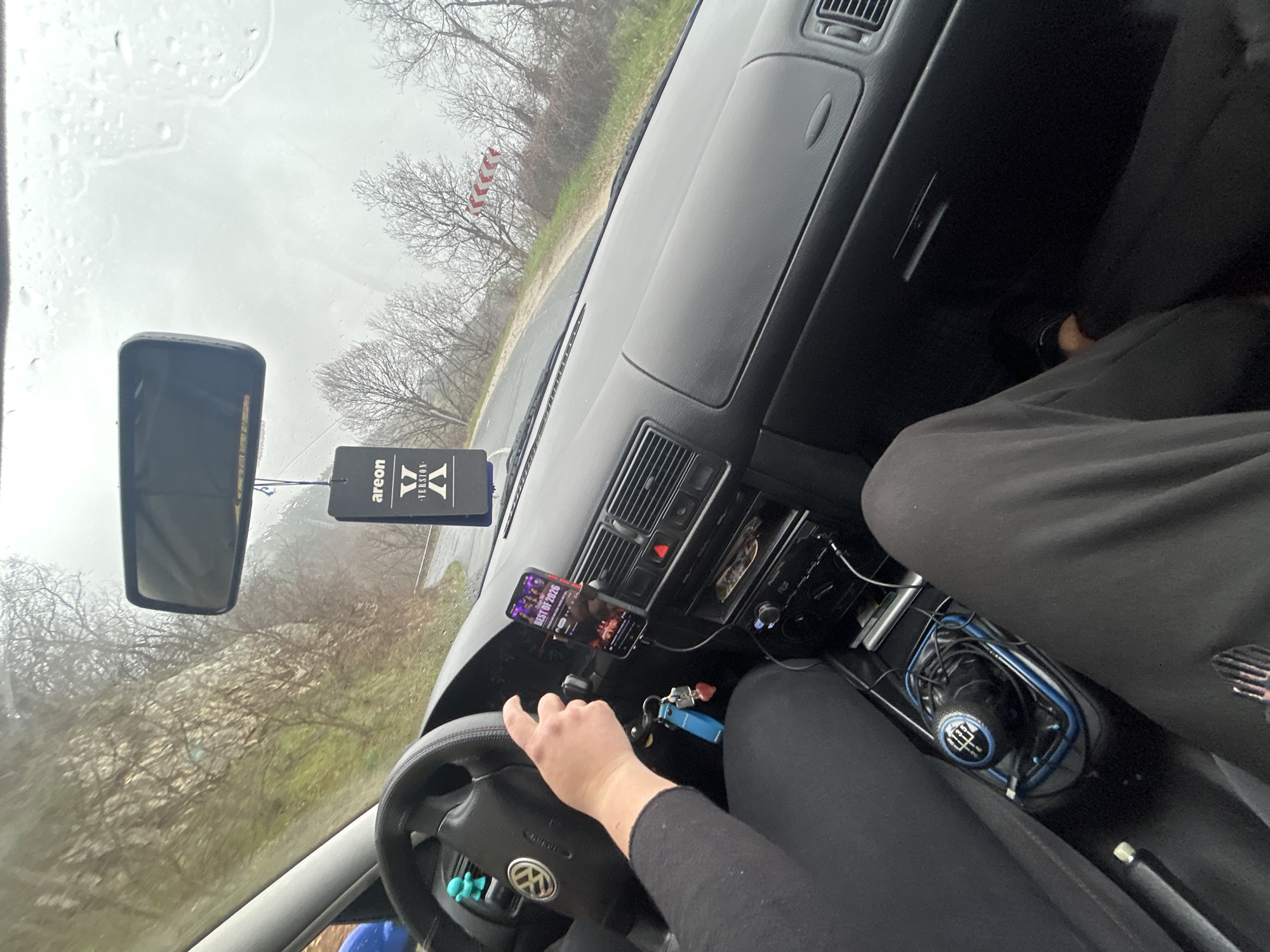 The scene is a view from inside a car, likely the driver's perspective. The windshield is wet, suggesting rain. A rearview mirror and an "Areon X" air freshener are hanging from the top of the windshield. The dashboard is visible with air vents and controls. A smartphone is mounted on the dashboard, displaying a music playlist titled "BEST OF 2026." Keys are hanging near the steering wheel, which has a Volkswagen logo. The driver's hand is on the steering wheel, and their legs are visible, clad in black clothing. The gear shift is also visible with blue accents. Outside the car, bare trees and a road can be seen. The presence of a chevron alignment marker suggests the location is on a curved road, potentially in the vicinity of Pechinska, Bulgaria, known for its winding roads and natural surroundings.