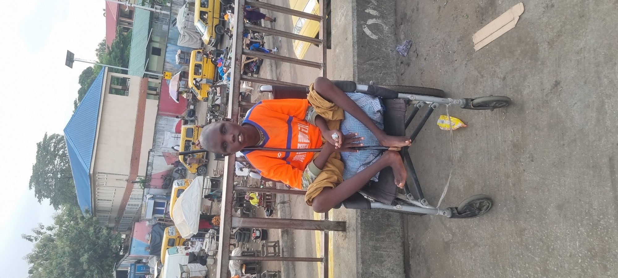 A young boy with dark skin sits in a wheelchair on a bridge in Lagos, Nigeria. He wears an orange t-shirt and tan shorts, his legs are crossed in the chair. The surrounding area is filled with yellow taxis and pedestrians, typical of a busy Lagos street. The bridge appears to be above a road or market area. Buildings with blue and green roofs are visible in the background. A bag of chips hangs on the side of the wheelchair. The surface of the bridge is concrete.