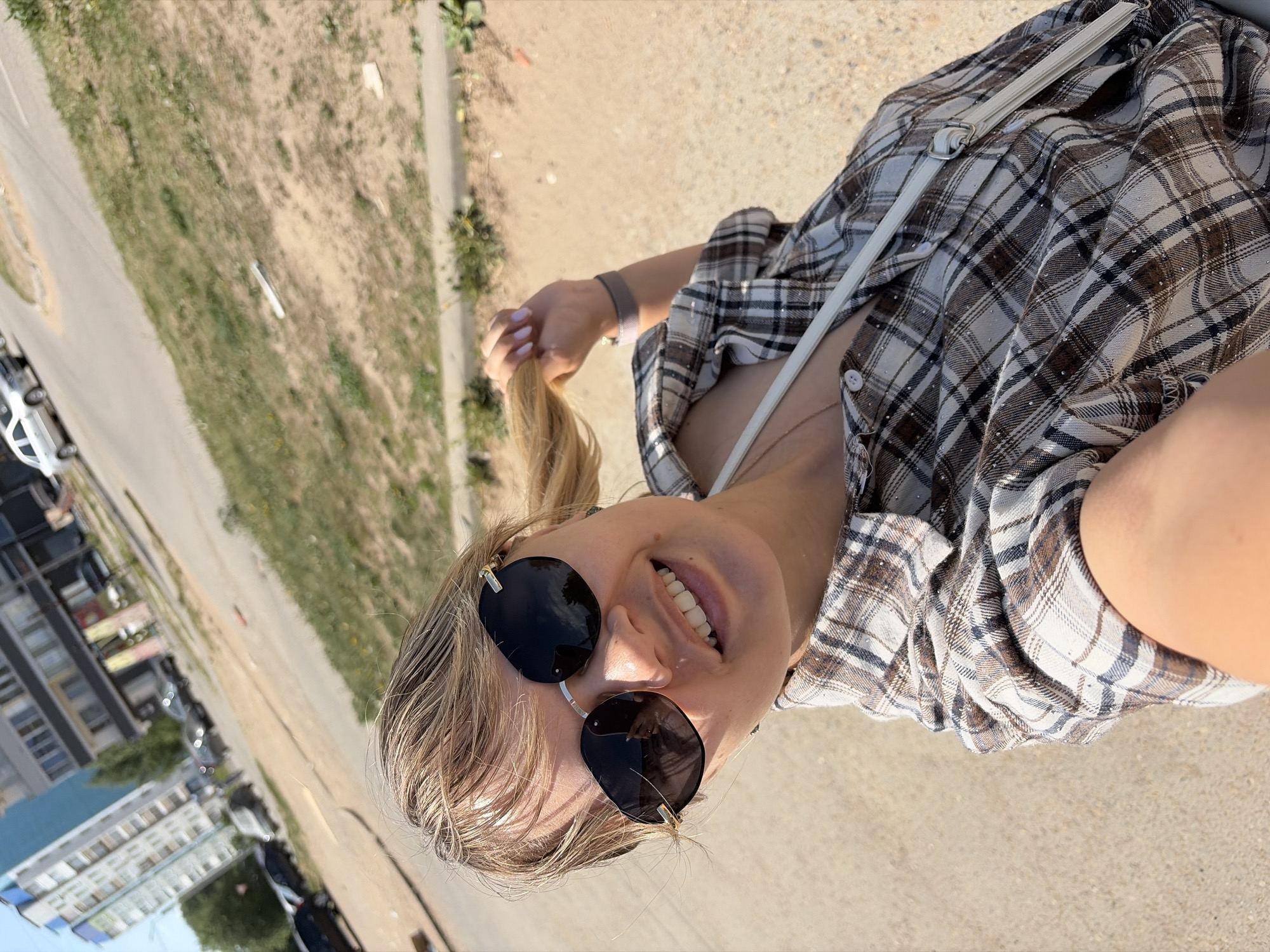 A vertically oriented, medium shot photograph captures a fair-skinned young woman with blonde hair and a nose ring, wearing round sunglasses. She is seen from a low angle and is smiling, facing toward the camera while outside. The background reveals a typical urban environment, located in the city of Chaikovsky, Russia. Visible in the background are buildings and vehicles parked along a street. Adjacent to the street is a grassy area, providing a contrast to the hard urban landscape. The woman is wearing a plaid shirt with a white strap from a bag over her shoulder. She is holding a strand of her hair in her hand. The lighting suggests it is daytime with bright sunlight.