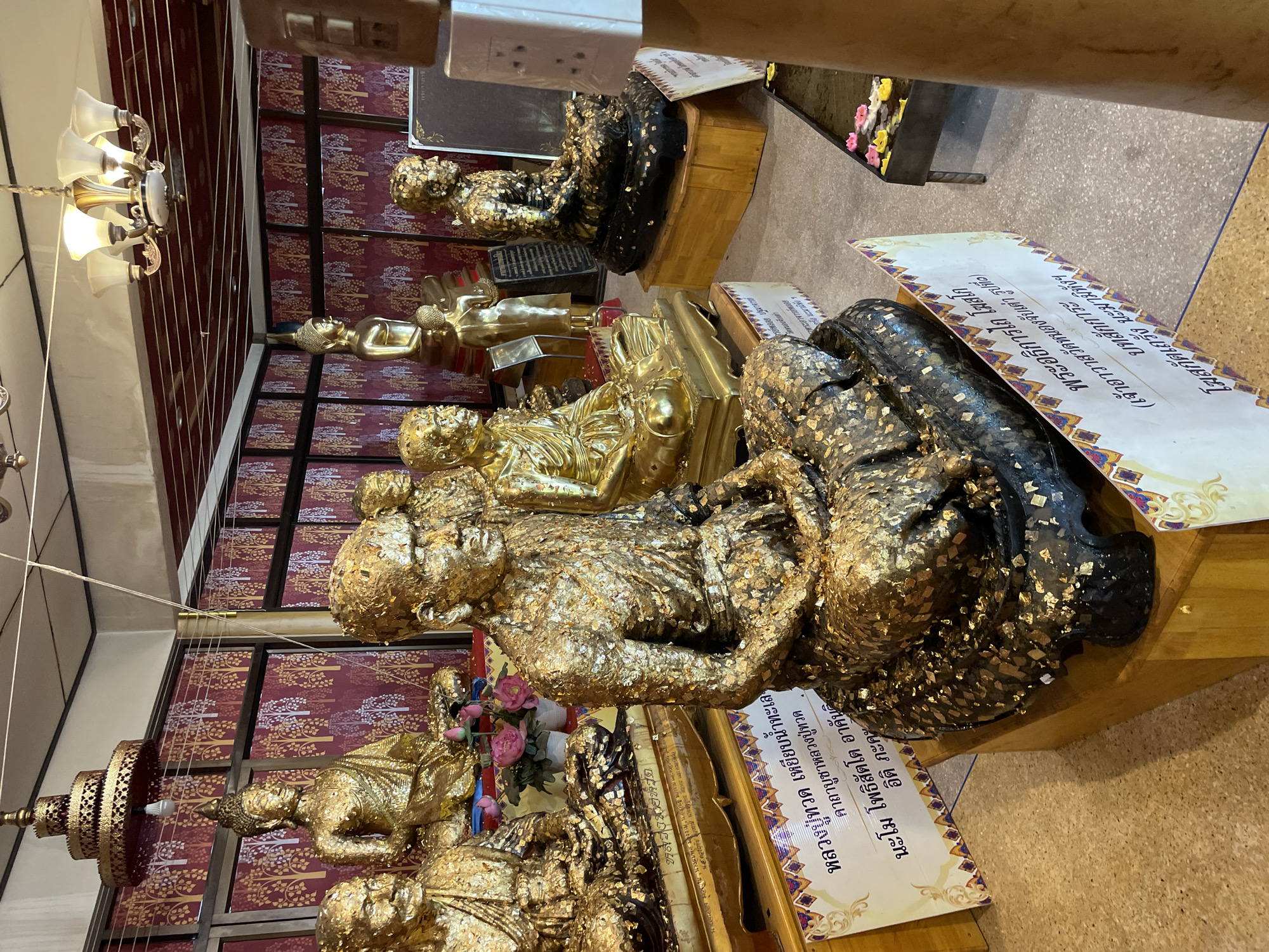 The image shows an indoor scene at a temple in Tambon Na Chom Thian, Thailand. The foreground is dominated by several gold-leaf-covered statues of Buddhist monks and a Buddha. The statues are seated in various meditative poses, some with their eyes closed and others with their eyes open. The gold leaf is applied unevenly, giving the statues a textured and worn appearance.
Behind the statues, a wall is decorated with a red patterned wallpaper featuring white and gold trees. A large golden chandelier hangs from the ceiling, casting a warm light on the scene. To the right, a small altar holds floating candles and some flowers. Several signs with Thai script are placed in front of the statues, likely providing information about them or religious offerings. The overall impression is one of reverence and tradition, with the abundant gold leaf signifying the sacredness of the site. The floor appears to be made of speckled concrete.