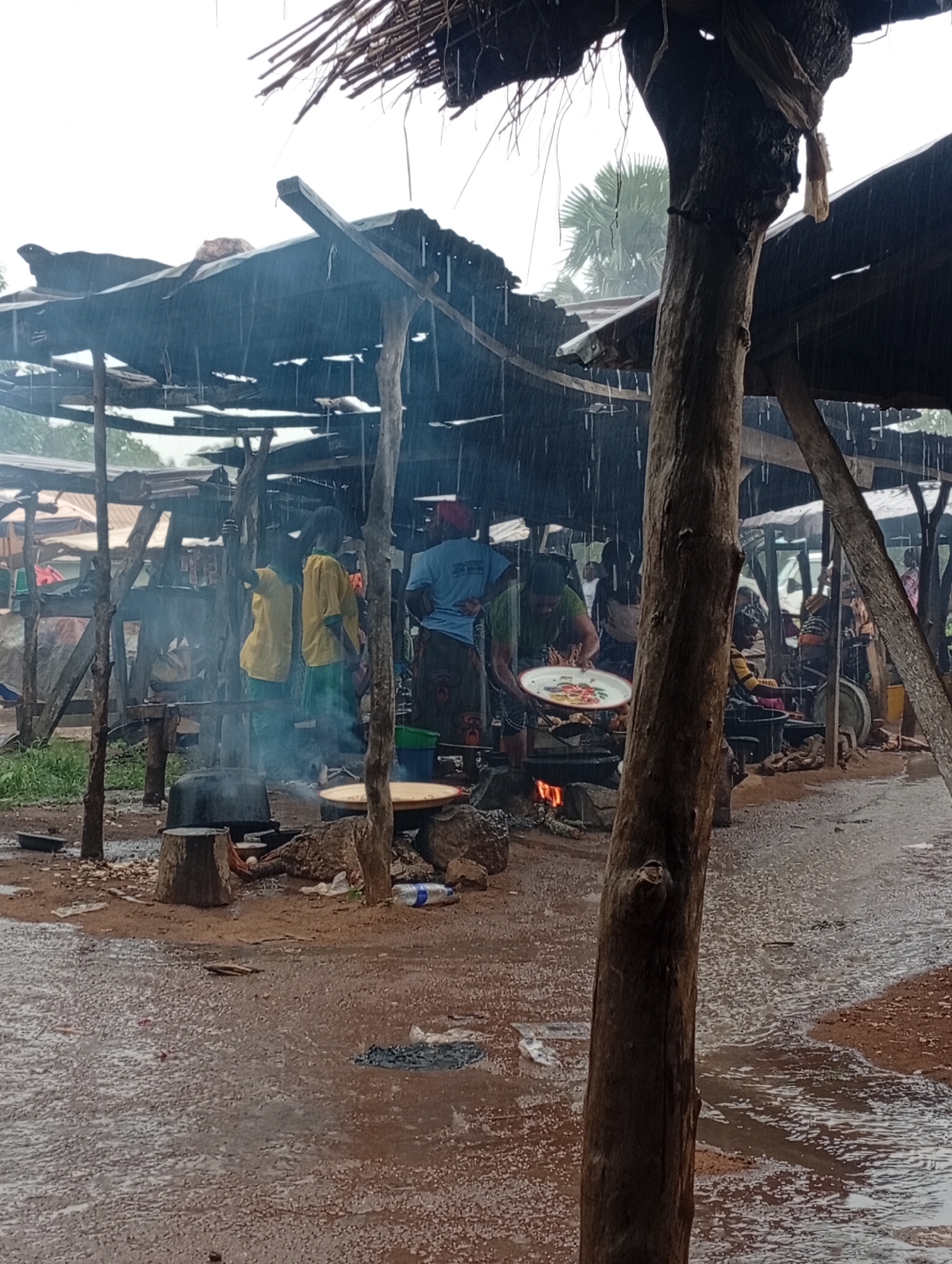In Andaha, Nigeria, during a heavy downpour, a bustling outdoor market scene unfolds. Several individuals, likely vendors and customers, are engaged in various activities under makeshift shelters constructed from corrugated metal and branches. Steam rises from cooking fires, indicating food preparation, with large pots and pans in use. One person is seen holding a large, decorated platter, possibly serving food. The ground is wet and muddy, with puddles forming from the rain, suggesting it is a rural or unpaved area. In the foreground, a large tree trunk stands prominently, and beyond it, palm trees are visible, typical of the Nigerian landscape. The overall atmosphere is one of resilience and activity despite the inclement weather.