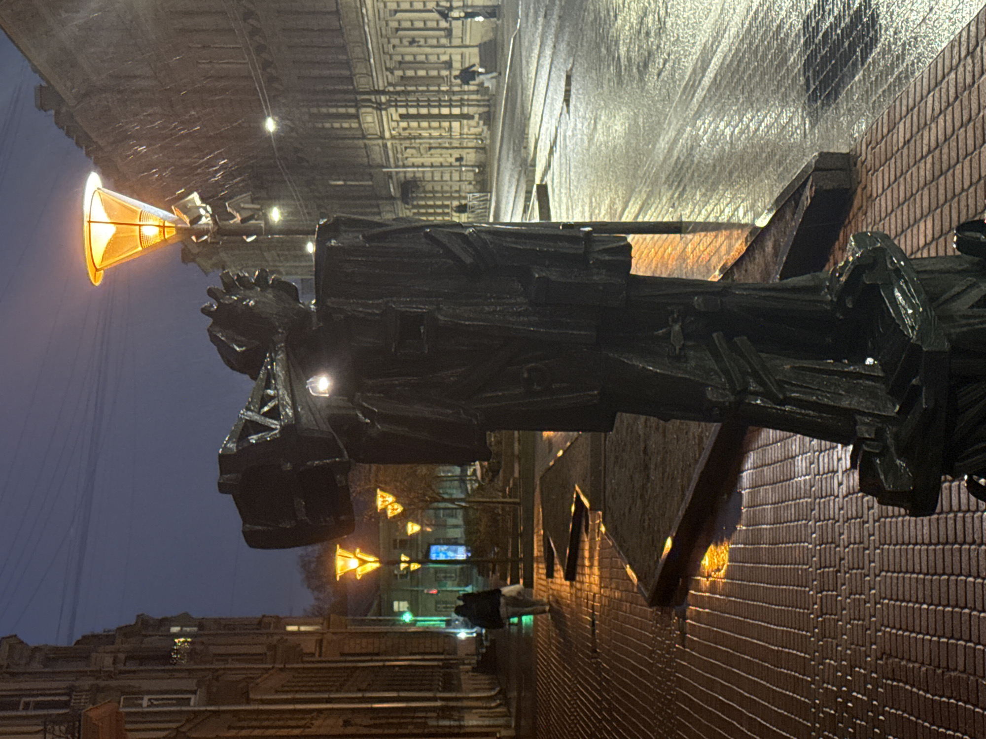 A medium-angle, rotated photograph depicts a monument to Nikolay Gogol in Saint Petersburg, Russia, viewed against a dark sky at dusk or night. The monument is made of dark, rough-hewn metal and appears to be elevated. The background features buildings and streetlights illuminating a wet, reflective surface. One illuminated streetlight has a distinctive cone shape. A person is visible walking on the wet street. The monument is mounted on a brick base.