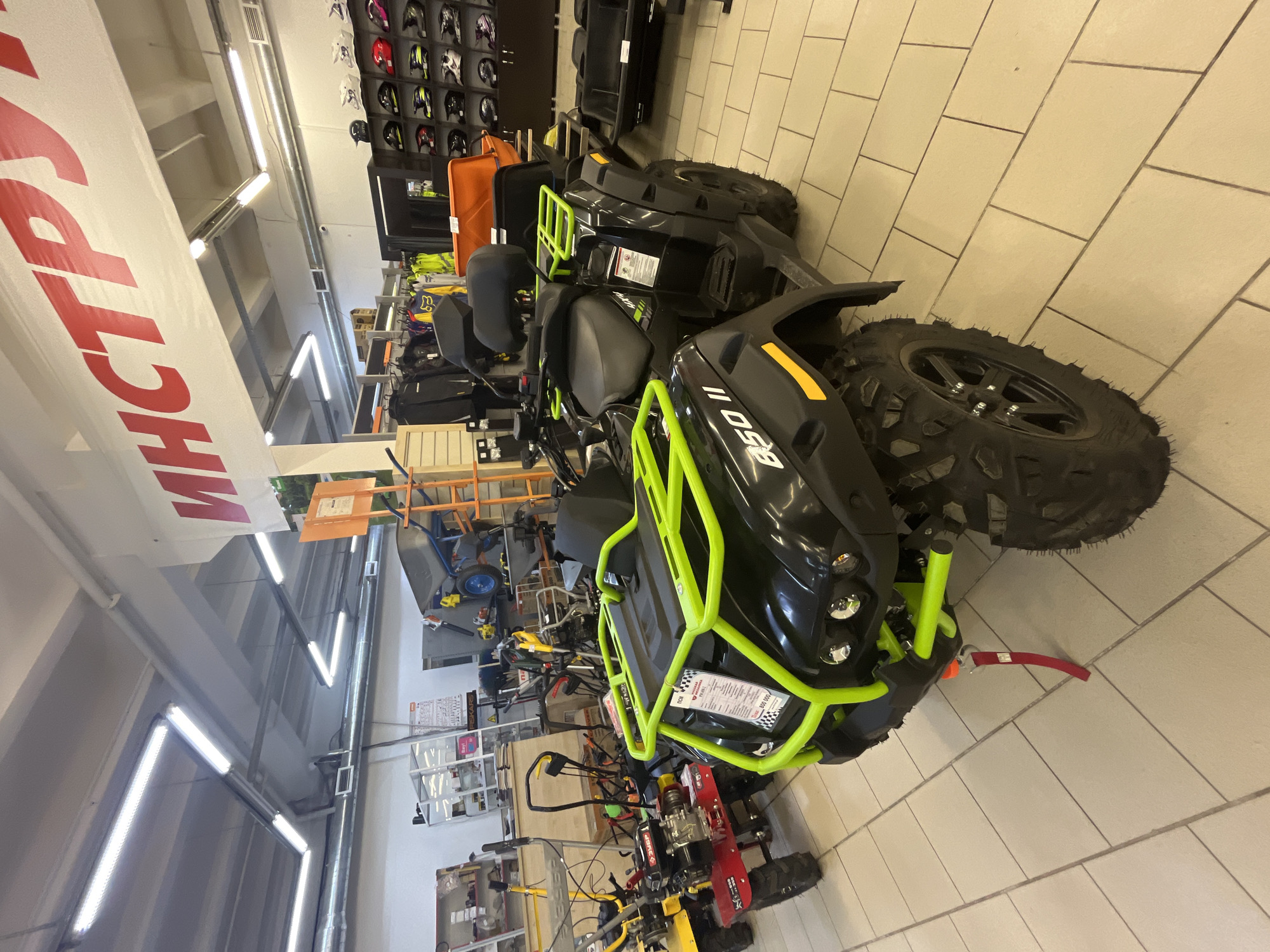 The image depicts the interior of a store in Chaikovsky, Russia, filled with various items including ATVs, possibly for sale. The store has bright overhead lighting. There are shelves stocked with merchandise, including motorcycle helmets, and other equipment such as tillers. A prominent banner with Russian text hangs from the ceiling. The floor is tiled. The perspective suggests the photo was taken from a standing position, looking down the aisle of the store.