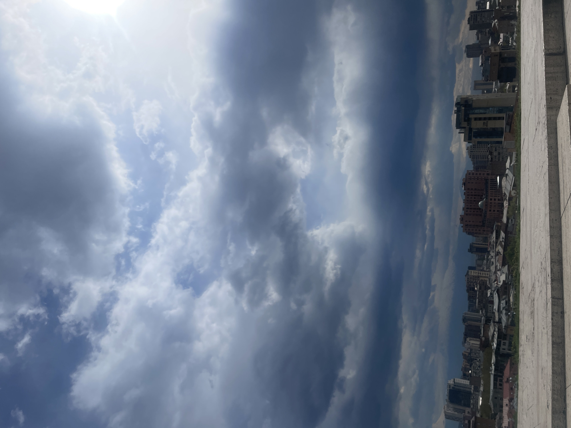 The image captures a scene in Yerevan, Armenia, showcasing a dramatic sky dominated by voluminous cumulus clouds, ranging from bright white to deep grey. The sun shines through the clouds, adding brightness to the upper portion of the frame. Below the sky, a cityscape is visible, with densely packed buildings of varying heights and architectural styles, indicating a mixed residential and commercial area. A light-colored, textured stone structure, possibly a wall or a part of a building, is present on the right side of the frame. No people are directly visible in the shot. The environment suggests a partly cloudy day in an urban setting.