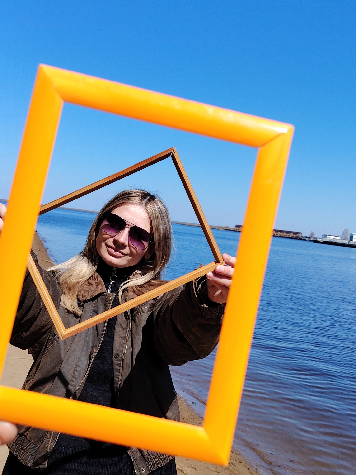 A woman stands on a sandy shoreline holding two square frames, one orange and one wood-toned, in front of her. She is wearing sunglasses, a black turtleneck sweater, and a brown jacket. The background features blue water and a clear blue sky. In the distance, there are buildings and structures along the shoreline. The location is Chaikovsky, Russia.
