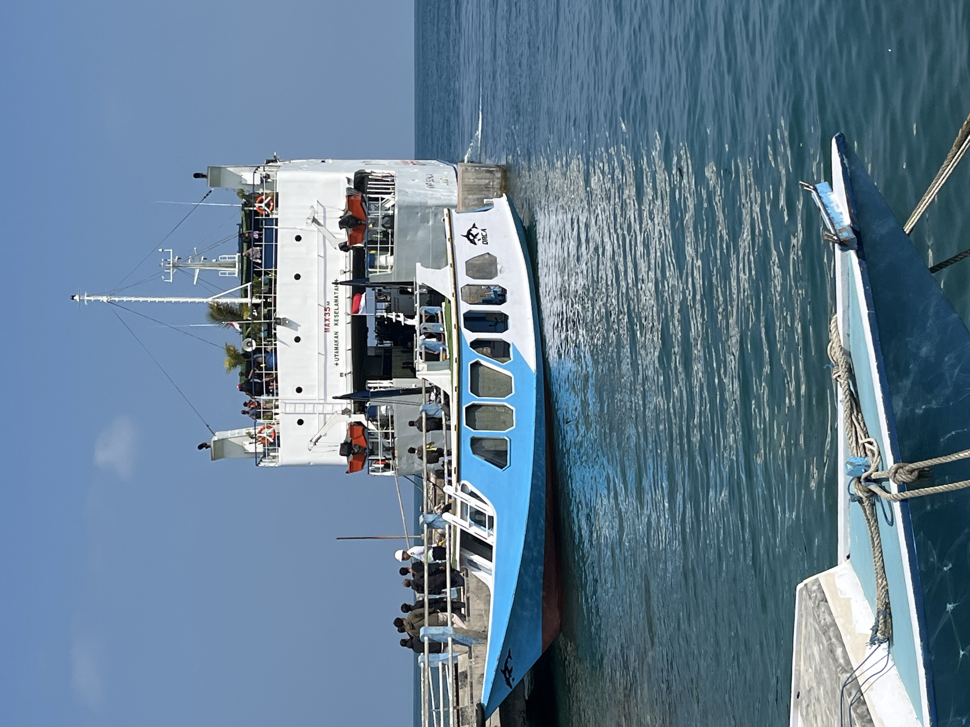The image depicts a bright, sunny day in the city of Karimunjawa, Indonesia, at a port. A large ferry, predominantly white with a blue hull, is docked and tethered to a pier. Several people are visible on the deck of the ferry and on the pier. A smaller, bright blue and white boat with a dolphin graphic on its bow is tied alongside the ferry, with its bow extending towards the foreground. The water is calm and reflects the sunlight. The sky is clear and blue. The overall scene suggests a busy port environment, possibly during a transfer of passengers or cargo.