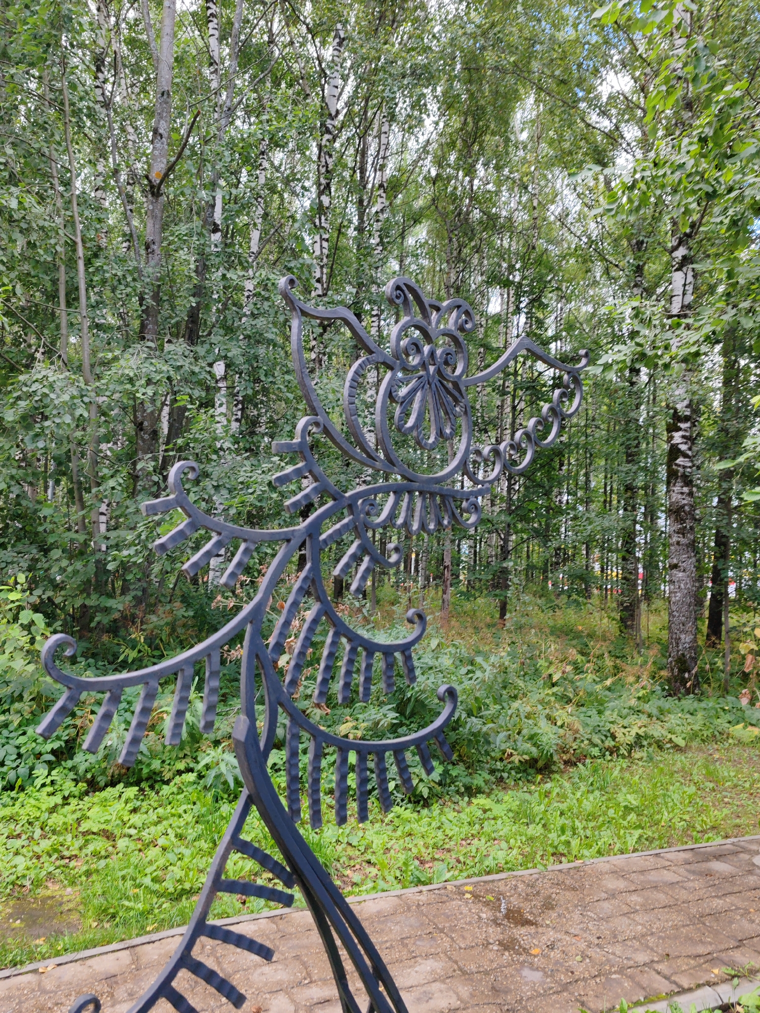 A dark gray, ornate metal sculpture resembling a bird or fantastical creature stands in the foreground of a wooded area with birch trees. The sculpture has intricate scrollwork and pointed feather-like elements. A paved walkway, possibly made of cobblestones or pavers, is visible in the lower portion of the image, with some wet patches suggesting recent rain. The background is filled with dense green foliage and the white trunks of birch trees, characteristic of a natural setting. The sculpture is positioned to the right of the frame, with the dense forest occupying the majority of the background. There are no people or animals visible in the image. This scene is located in the Central Park of Culture and Recreation of Syktyvkar, Russia.