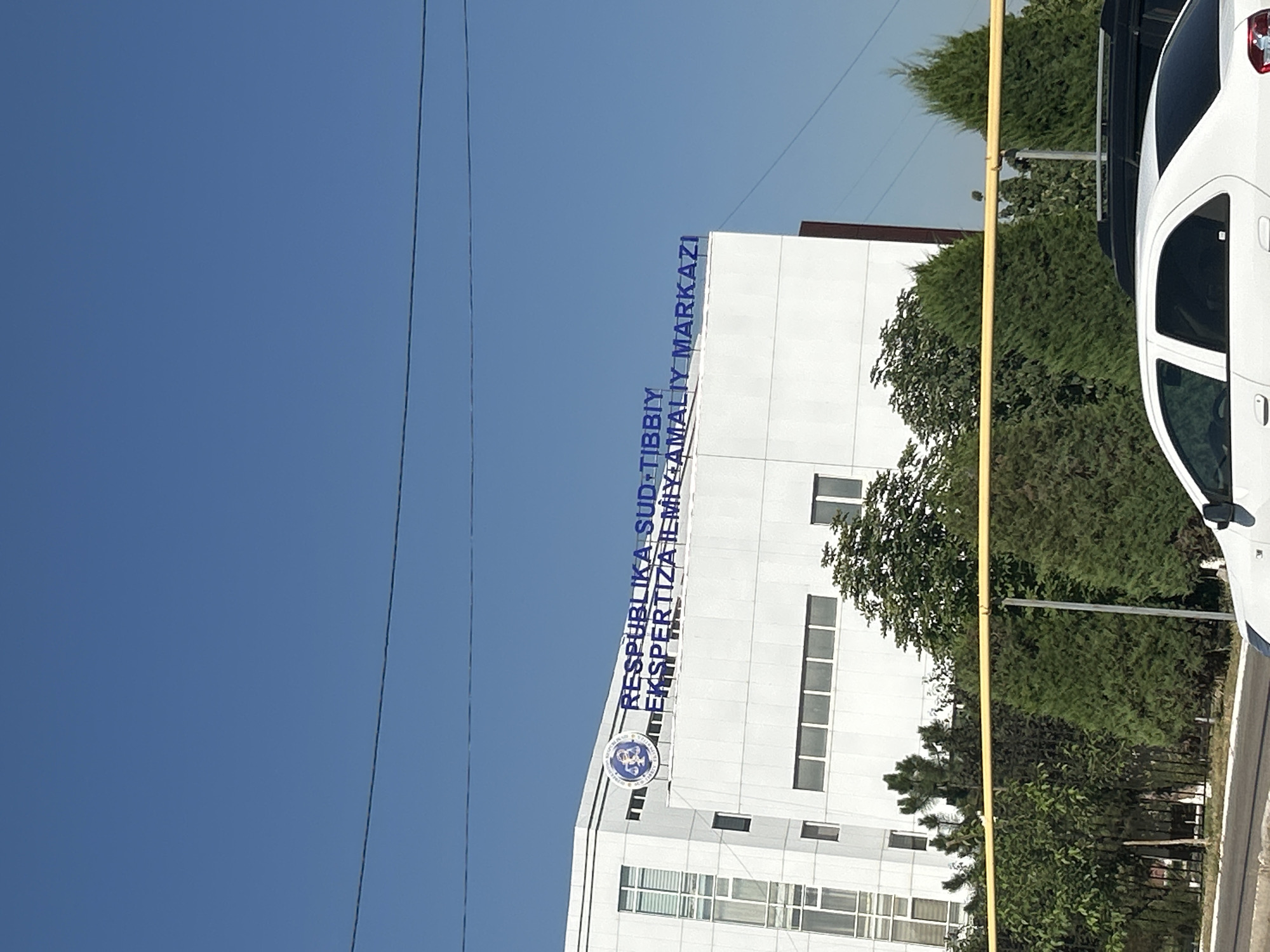 A white, multi-story building with a sign in blue Cyrillic letters stands against a clear blue sky. The sign reads "RESPUBLIKA SUD-TIBBIY EKSPERTIZA ILMIY-AMALIY MARKAZI" and is accompanied by a circular logo with an eagle. The building has a modern architectural style with large windows. In front of the building, there are several trees, a black metal fence, and a yellow pipe running horizontally. A white car is partially visible in the foreground, suggesting a street or parking area. The scene appears to be a sunny day in a city environment. This is the building of the Republican Scientific Practical Center of Forensic Examination in Tashkent, Uzbekistan.