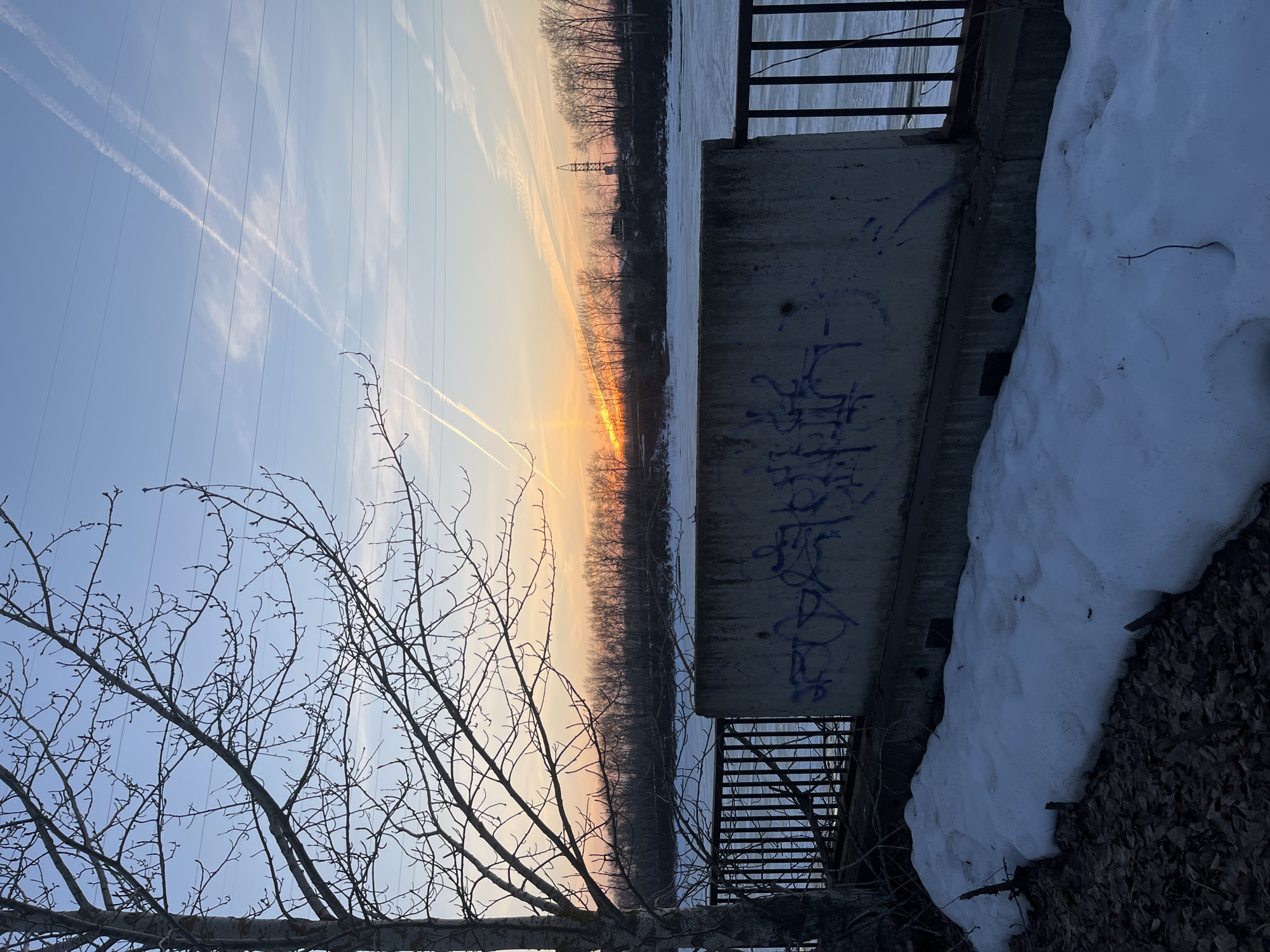 The image shows a sunset over a partially frozen river in what appears to be a slightly industrialized area. Contrails are visible in the sky, and power lines run across the horizon. A concrete structure with graffiti and a metal railing is in the foreground, along with a patch of snow and bare branches. Without further information, I cannot confirm the specific location within Chaikovsky, Russia.