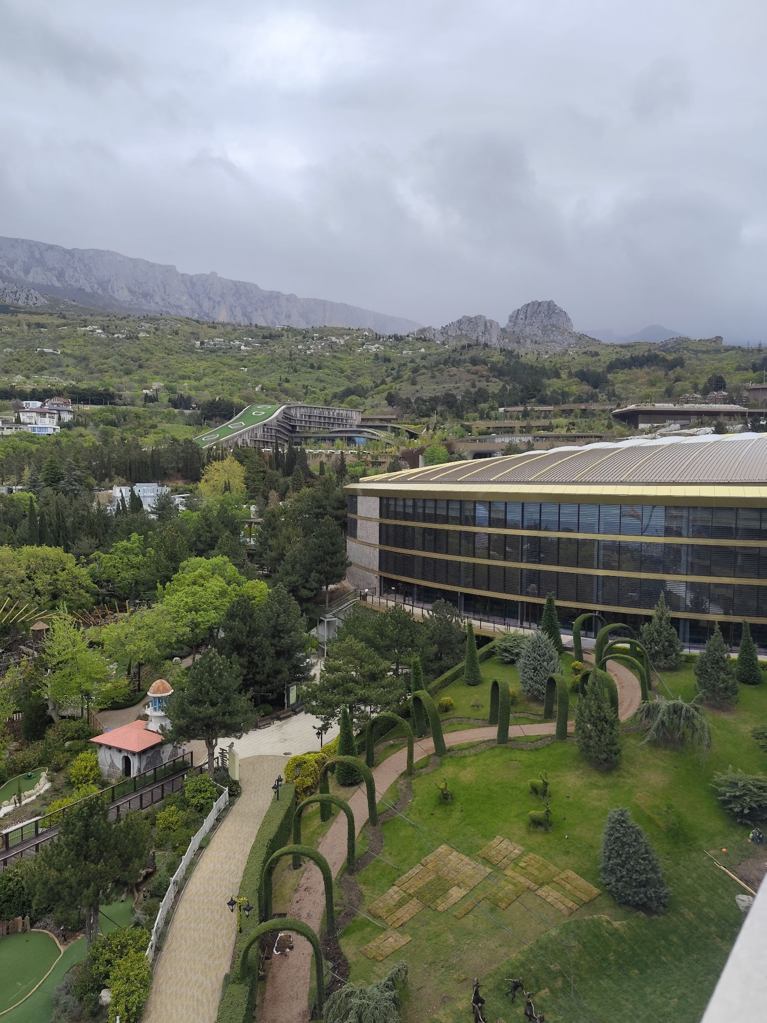 The image shows an elevated view of a section of Симеизский поселковый совет, Ukraine on a cloudy day.
A large, modern building with many windows occupies the right side of the frame. A garden and pathway featuring meticulously trimmed hedges and trees is situated in front of it. The pathway is lined with small, arched hedges.
In the foreground is a pathway with a mini-golf course on the left. There is a small, decorative building along the pathway.
In the background are hills covered in green vegetation and scattered buildings. A mountain range is visible in the distance, obscured by clouds. The overall atmosphere is overcast.