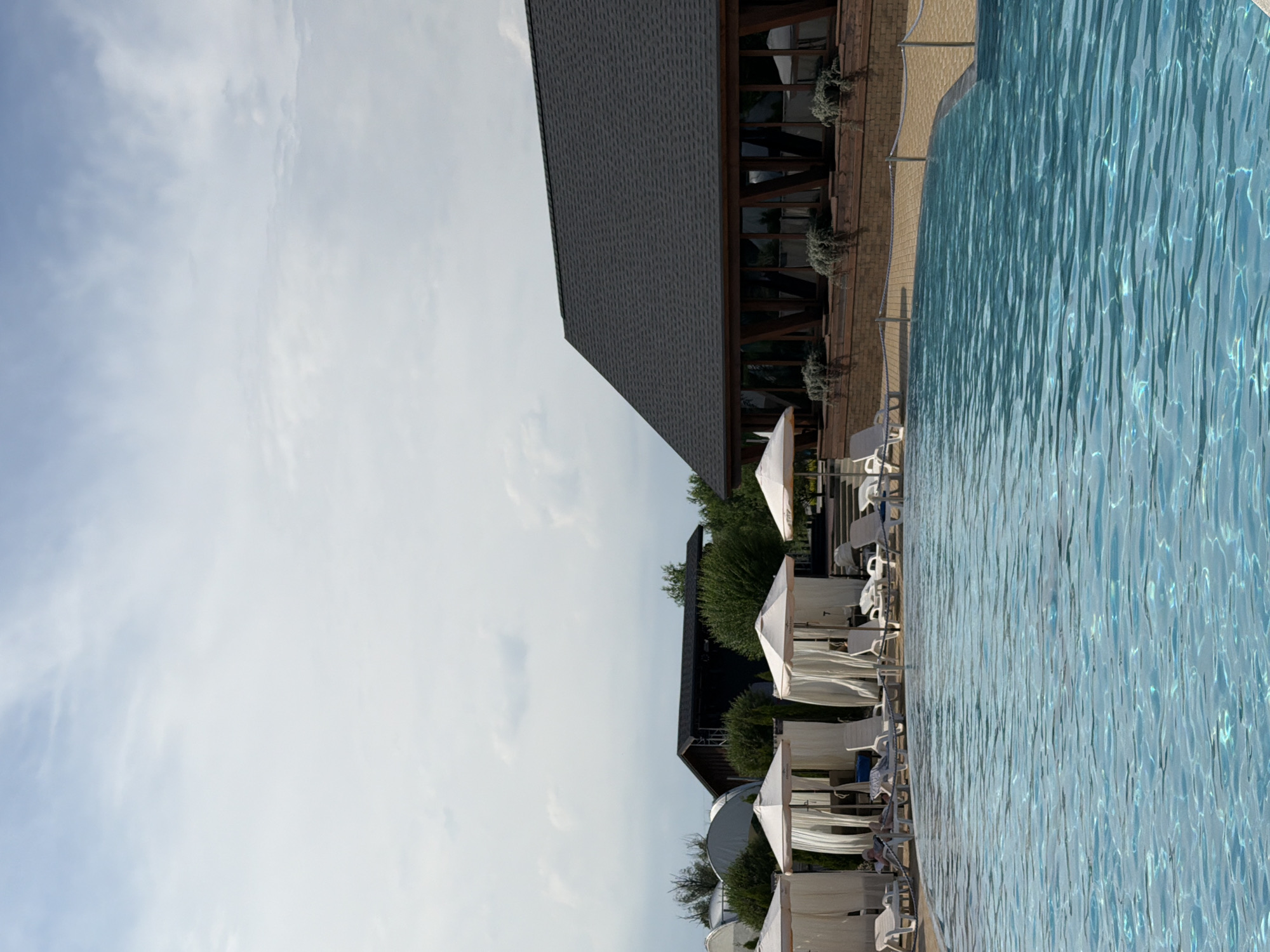 The image depicts an outdoor swimming pool area at what appears to be a resort or hotel, possibly located in Kamyshin, Russia. In the foreground, a large portion of the bright blue swimming pool is visible, with gentle ripples on its surface. Along the edge of the pool, there are several white lounge chairs and cabana-style seating areas, some of which are occupied by individuals. White umbrellas and curtains adorn these shaded resting spots.
In the background, a dark-roofed building with a modern architectural style featuring large windows and wooden accents stands prominently. Surrounding the building are green trees and other structures, including what seem to be white tents or covered areas. The sky above is partly cloudy, with patches of blue sky visible through the white clouds. The overall scene suggests a tranquil, recreational setting, likely during daylight hours.