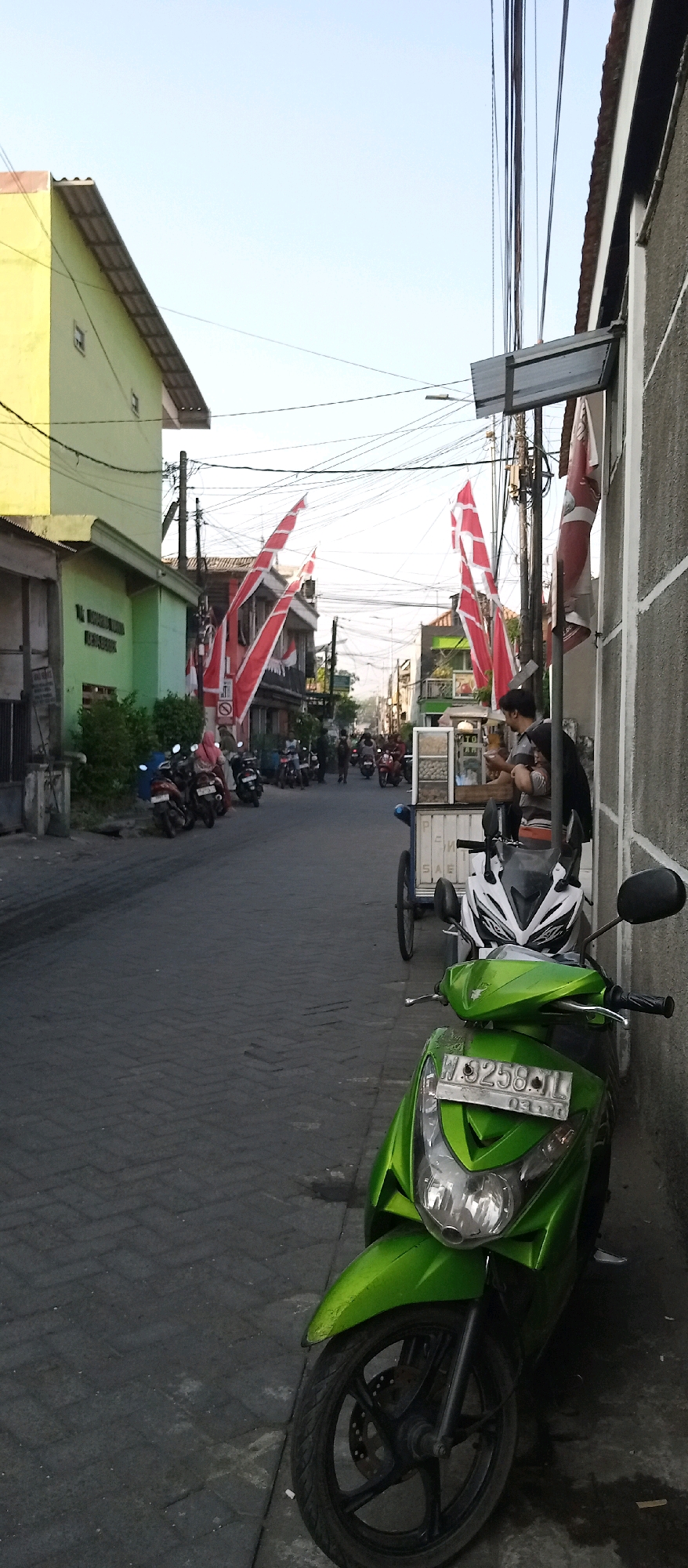 The image depicts a street scene in Wadungasri, Indonesia, with several motorcycles parked along the side and others in motion. Red and white flags, likely representing the Indonesian national flag, are displayed prominently on buildings and poles. In the foreground, a bright green scooter is parked. Further down the street, a food cart is visible, with two individuals standing near it. One person appears to be serving food, while the other is looking on. Buildings, some painted in bright colors like yellow and green, line the street. Utility wires are strung across the sky. The street is paved with brick-like material. The sky is clear and bright, suggesting it is daytime.