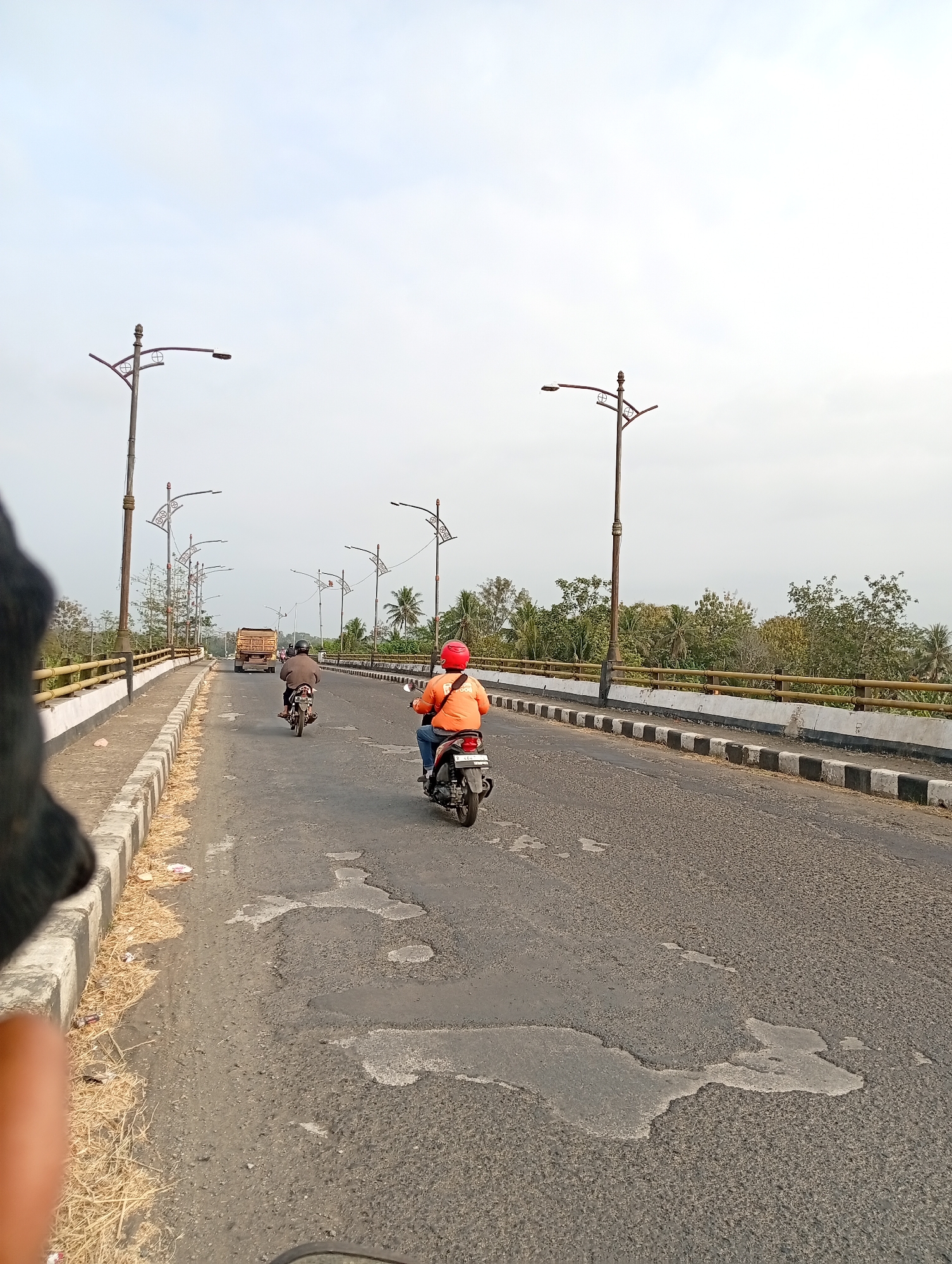 An eye-level, medium shot captures a view from the perspective of a vehicle traveling over a bridge in an outdoor, daytime scene. Two motorcyclists are visible on the asphalt road. The lead motorcyclist, wearing a red helmet and an orange shirt, is on a scooter. Behind them, another motorcyclist is riding a scooter, wearing a brown jacket and a helmet. A truck is seen in the distance. The road is lined with decorative lampposts on both sides. A bridge railing, composed of a yellow metal bar and white concrete posts with black and white stripes at the bottom, runs along the side of the road. The environment is characterized by trees and foliage beyond the railing, and the sky is overcast. The road surface shows signs of wear and tear, with patches of damaged asphalt. The left side of the frame is partially obscured by a dark object, possibly part of the capturing vehicle.