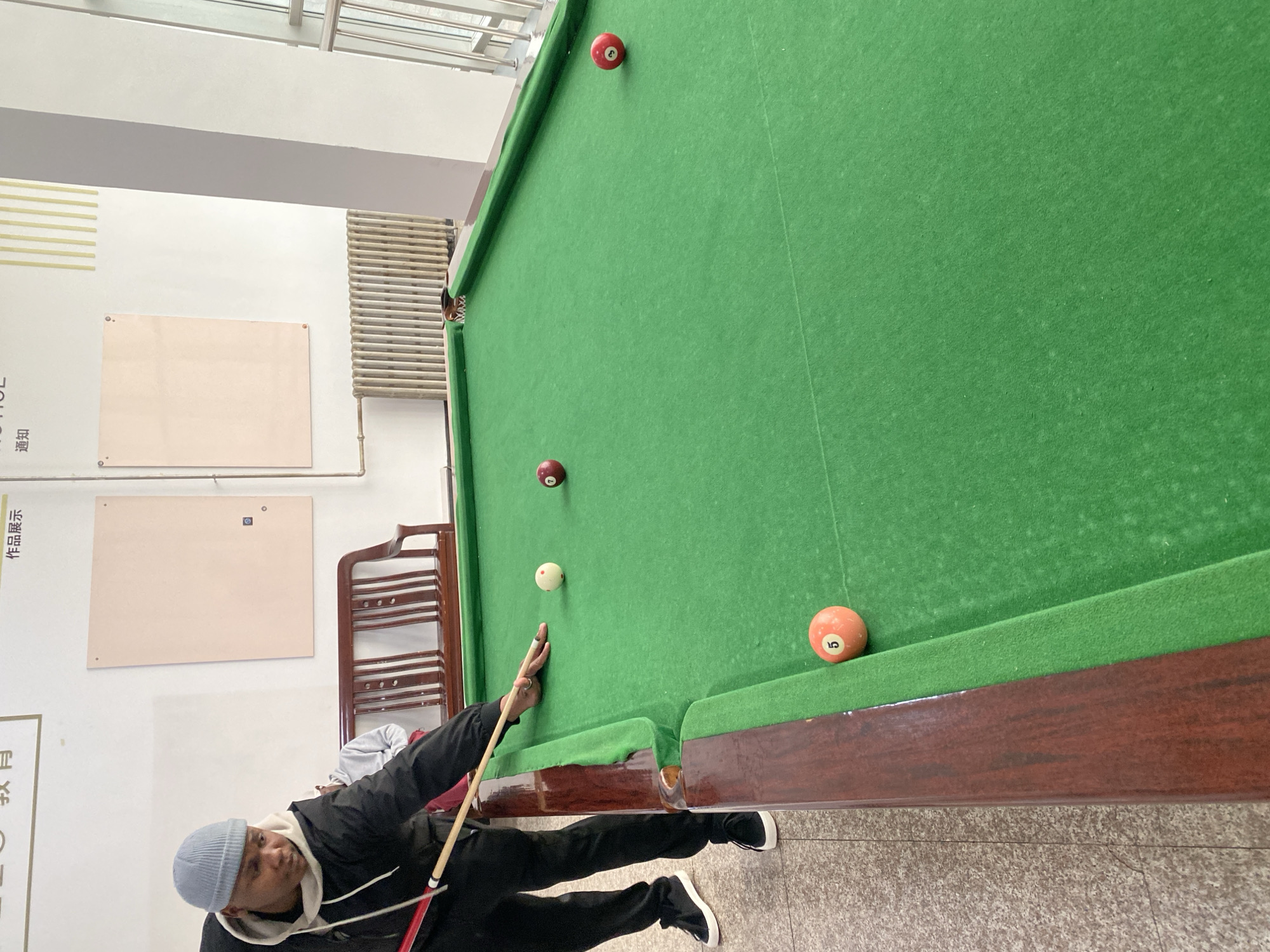 A man in a blue beanie and black jacket is playing pool indoors. He is holding a pool cue, poised to strike a white ball on a green pool table. Two red pool balls are also visible on the table. One red ball is near the upper left corner pocket, and the other is near the center. An orange ball with the number "5" is near the lower right corner. The pool table is surrounded by a wooden frame, and the floor is tiled. In the background, there are two bulletin boards on a white wall. A radiator is visible near the pool table. The location is Zhongtun, China.