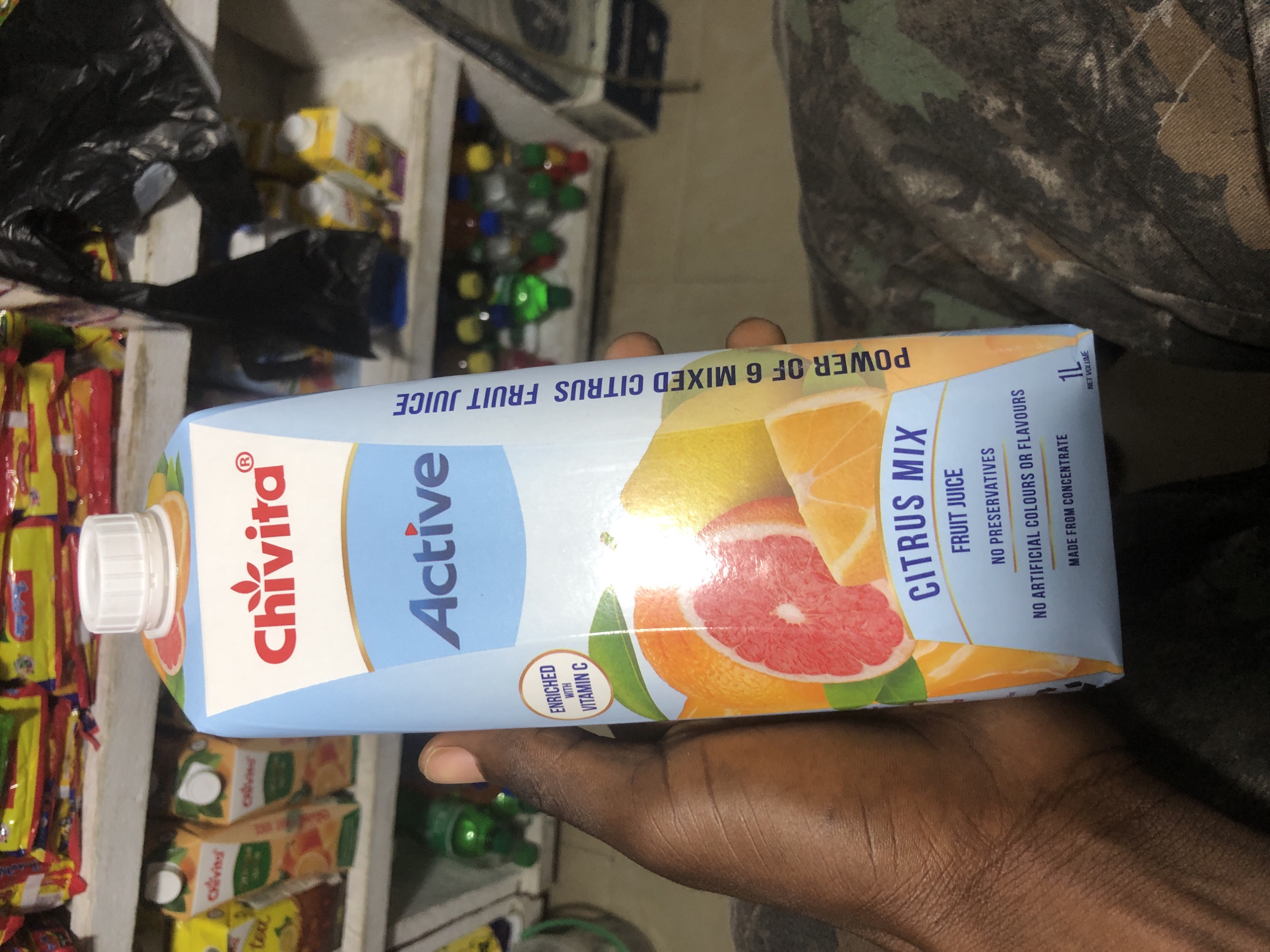 A hand holds a carton of Chivita Active Citrus Mix fruit juice. The carton is light blue with white and orange accents. The brand name "Chivita" is in red letters at the top, with "Active" in large blue letters below. The carton displays images of various citrus fruits like oranges and grapefruits. Text on the carton indicates it is a "FRUIT JUICE," "NO PRESERVATIVES," "NO ARTIFICIAL COLOURS OR FLAVOURS," and "MADE FROM CONCENTRATE." The net volume is listed as 1L. The background shows shelves stocked with other food items and bottles, suggesting a retail environment like a shop or supermarket. The image was taken in Ado, Nigeria.