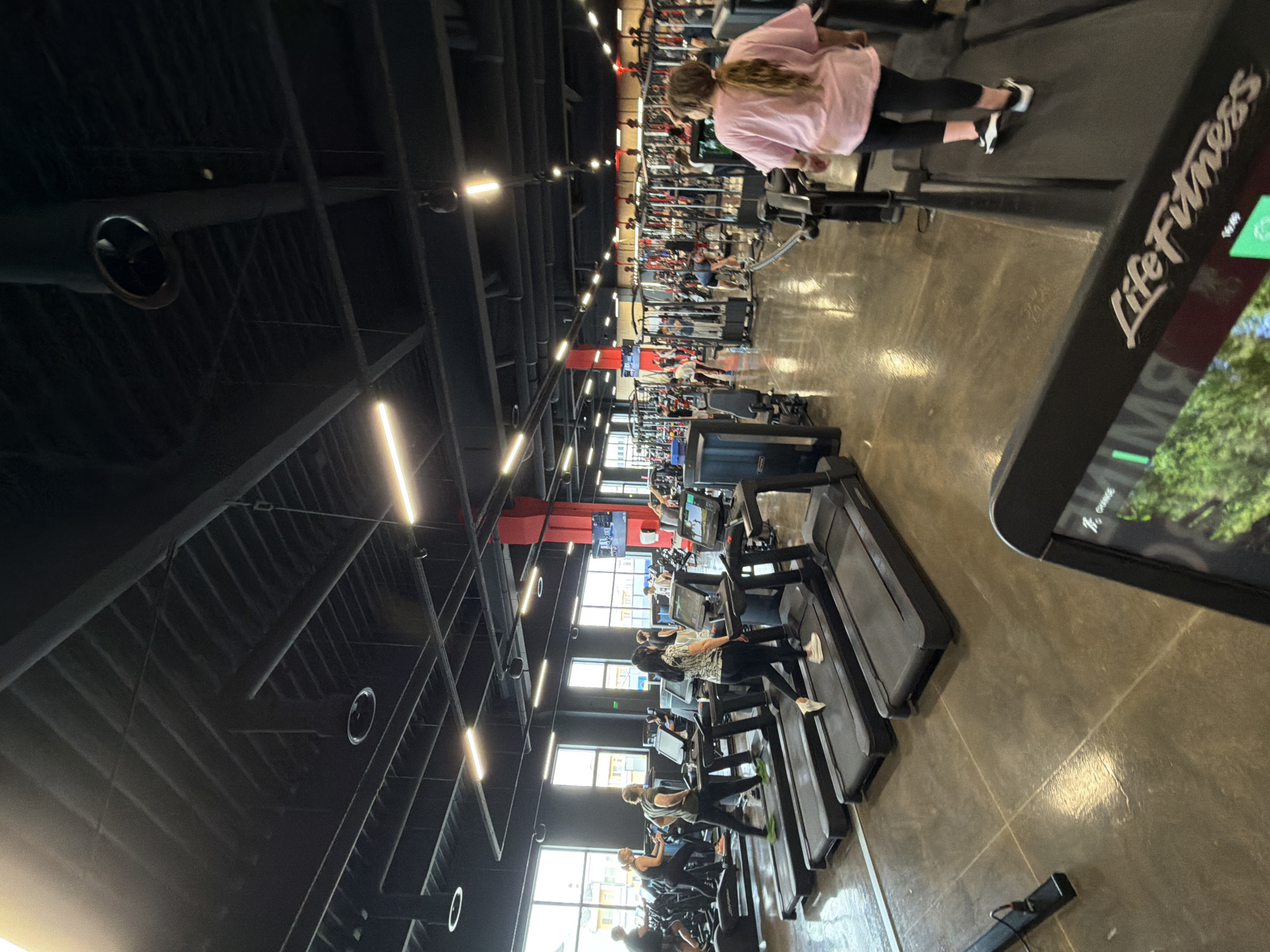 The image depicts the interior of a Life Fitness gym. The space is large, with high ceilings and visible structural supports. Artificial lighting is provided by linear fixtures. The gym is populated with various exercise machines, including treadmills, which are occupied by several individuals, some of whom are actively exercising. The treadmill in the foreground is displaying a Life Fitness logo and a visual interface. The scene suggests a bustling gym environment. Location cannot be determined.