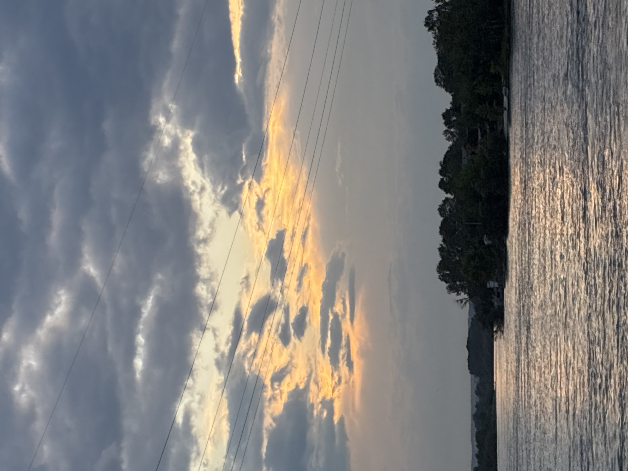 The image shows a body of water, likely a river, in Irkutsk, Russia, during what appears to be sunset. The sky is filled with dramatic, dark gray clouds interspersed with streaks of golden light from the setting sun. Several power lines are visible in the upper portion of the frame, stretching horizontally across the sky. The water's surface reflects the muted light of the sky, creating a shimmering effect. Along the far bank of the water, a line of dense green trees forms a silhouette against the horizon. There are no people or specific identifiable landmarks visible in the image that would pinpoint a precise location within Irkutsk. The scene is tranquil and evocative of a natural landscape.