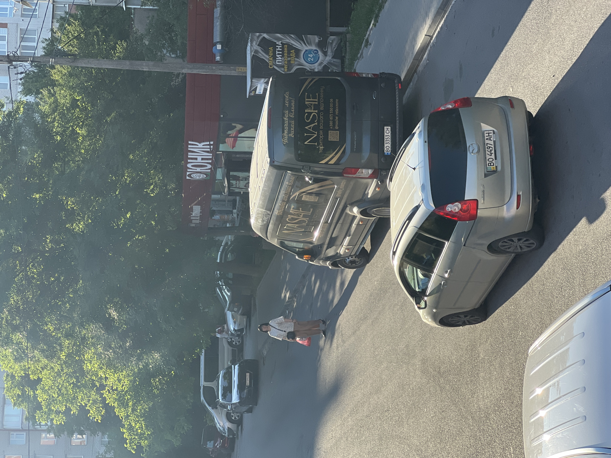 The image captures a street scene in Ternopil, Ukraine. In the foreground, the rear of a silver Mazda 5 wagon is visible, parked on the right side of a paved road. The car displays the license plate BO 4497 AH. Further up the road, a dark gray Ford Transit van, identified by the license plate BO 3353 CH, is parked on the right. The van has the branding "NASME" in large gold lettering, along with Ukrainian text. A woman in a light-colored blouse and dark pants is standing on the sidewalk to the left of the van, holding a red plastic bag. Several other vehicles are parked along the street, including a silver minivan and a dark-colored sedan. In the background, a red building with the word "ЮНИК" is visible, and beyond that, a multi-story residential building. Lush green trees with dappled sunlight filter through the scene. The overall impression is that of a typical sunny day in a Ukrainian city.