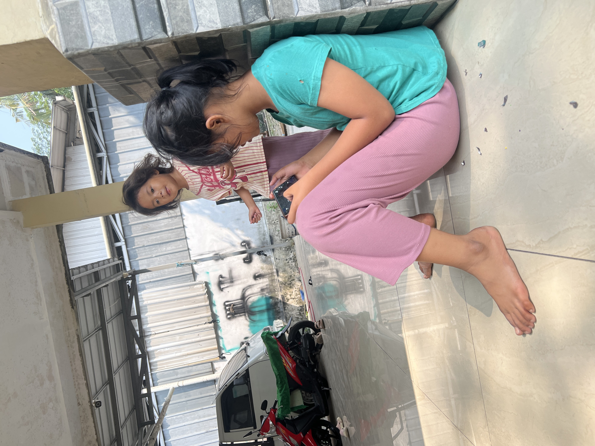 Two young girls are in an outdoor, covered area, likely a garage or carport, based on the presence of a white van and a red motorcycle. The girl in the foreground, with her back to the camera and facing right, is sitting on the tiled floor, wearing a teal t-shirt and pink pants. She is looking down at a dark-colored smartphone in her hands. Her dark hair is pulled back in a ponytail. To her left and slightly behind her stands a younger girl, facing the camera and looking towards the right. She has dark, wavy hair and is wearing a striped t-shirt with red and white stripes and a red collar, and a light purple skirt. Her left hand is extended slightly. The floor is tiled with a beige and cream pattern. In the background, the white van is parked to the left, and the red motorcycle, covered with a green cloth, is in front of it. The wall behind the van has graffiti art. The environment suggests a sunny day, with shadows cast on the floor. The general setting appears to be residential.
