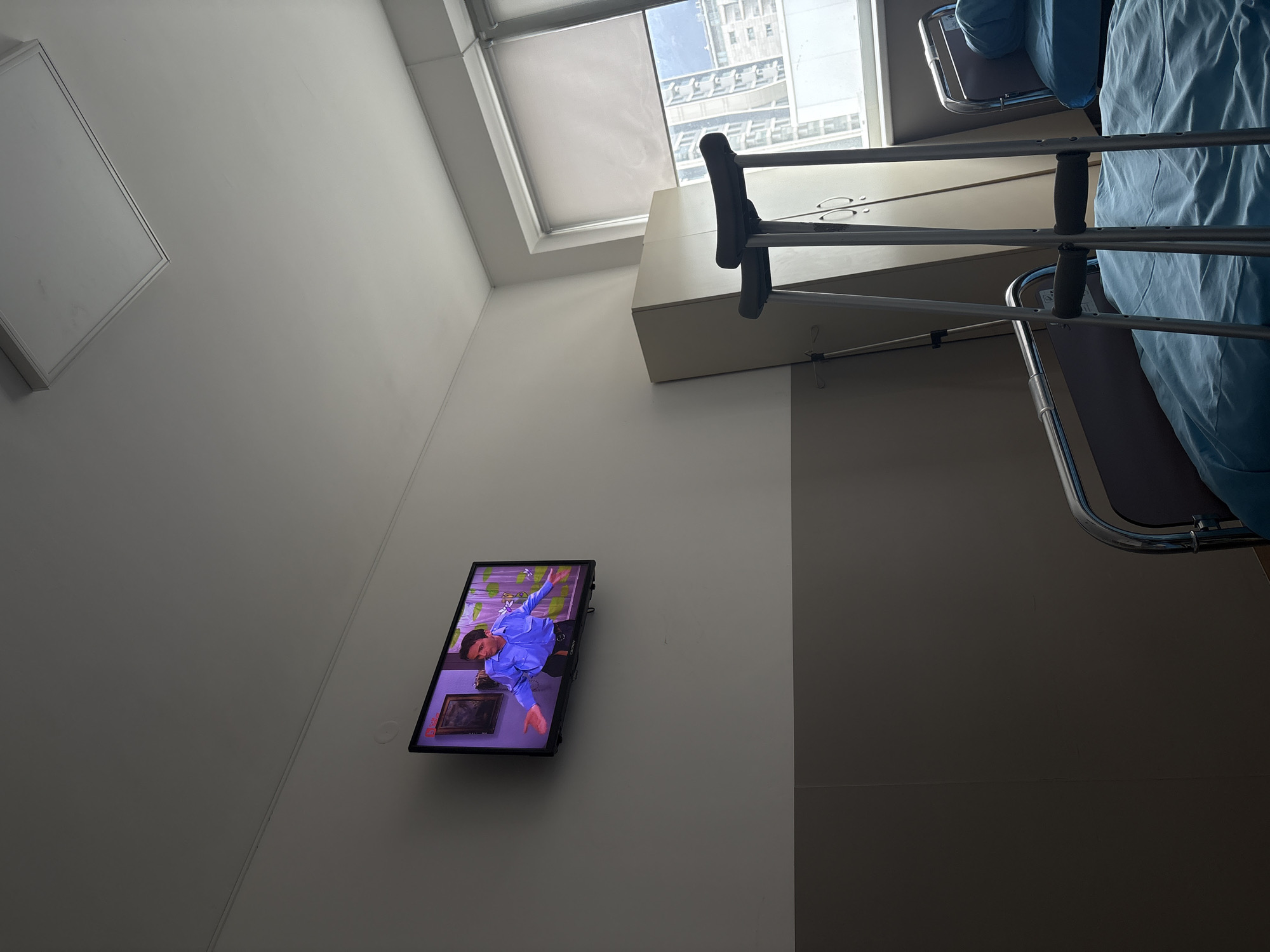 The image shows the interior of a hospital room. A patient's bed with blue sheets is visible on the right, with a pair of crutches leaning against it. A television is mounted on the wall, displaying a scene with a man in a blue shirt. A window provides natural light, revealing buildings outside. The location is Sofia, Bulgaria.