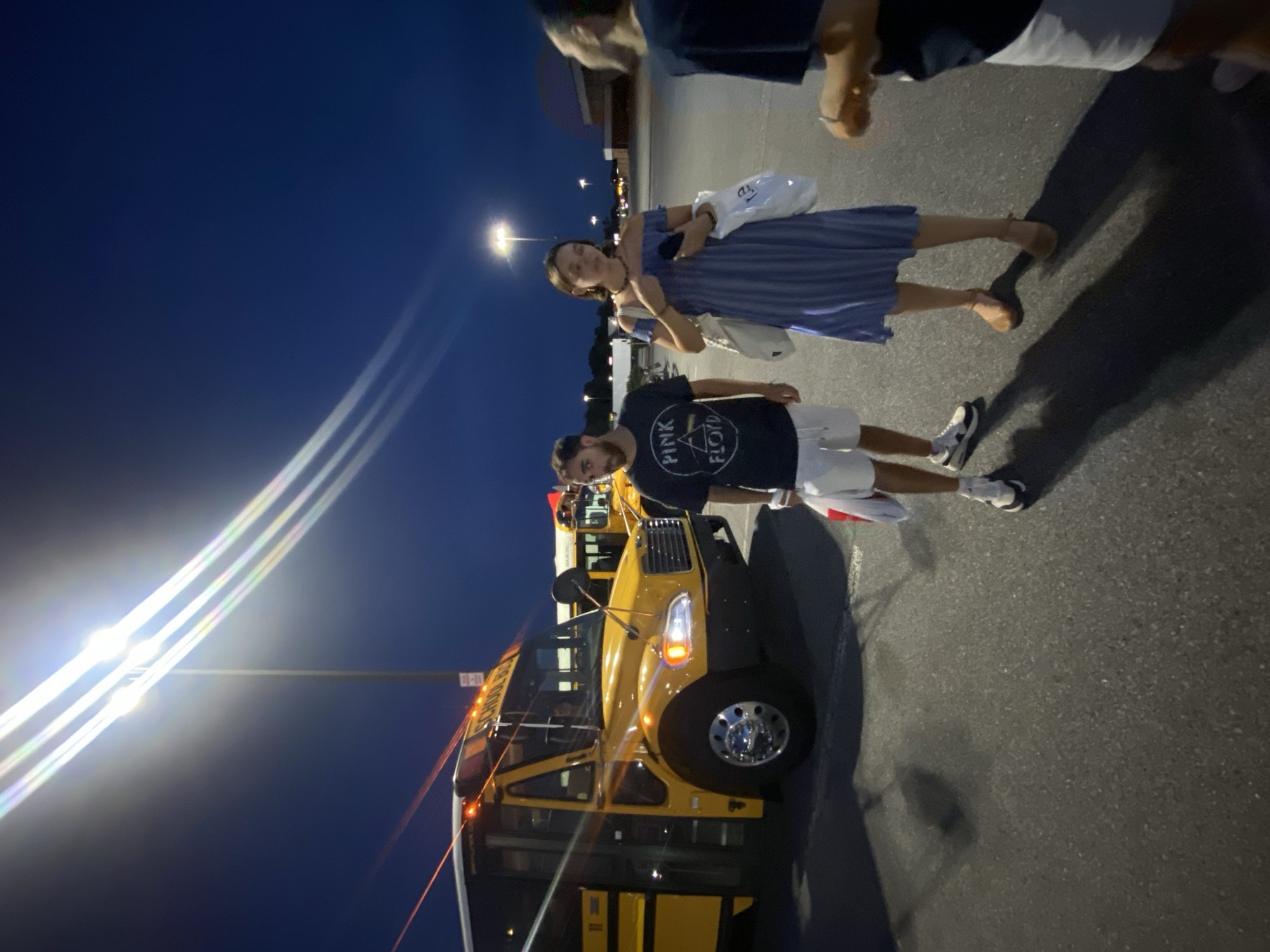 A group of four people are standing in a parking lot at dusk, near a yellow school bus. The sky is a deep blue, with streetlights casting bright beams and lens flares. One person, a man with dark hair, is wearing a black t-shirt with a "Pink Floyd" logo and white shorts, and he appears to be walking towards the viewer. To his right, a woman with dark hair is wearing a blue and white striped off-the-shoulder dress and brown sandals, carrying two white tote bags. Another person, whose back is to the camera, is wearing a dark t-shirt and white shorts, and is standing to the right of the woman. A fourth person, partially visible on the far right, is wearing a dark t-shirt. The school bus is the primary focus of the left side of the image, with its lights on. Another school bus is visible in the background, further back. The ground is asphalt, and shadows are cast by the people and the bus. The scene appears to be set in the city of Halfway, United States of America.