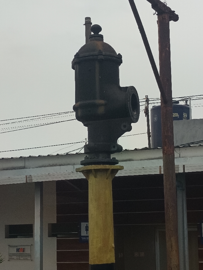A weathered dark industrial valve, featuring a domed top with a small finial and an exposed flanged opening on its right side, stands mounted on a yellow columnar pedestal with a flared capital and a black base. Behind this structure, a single-story building with a light-colored roof and dark walls is visible. A white sign with the red KAI (Kereta Api Indonesia) logo is affixed to the building's left facade. To the right of the yellow pedestal, a "Toilet W" sign with a female pictogram is visible on the building wall. Overhead, multiple black electrical wires traverse the scene. A rusty, reddish-brown vertical pole stands to the right of the main subject. Further in the background, a dark blue water tank bearing "DAL" and "SNI" markings is visible on a rooftop. The sky is uniformly overcast and grey. No people are visible. The scene is located at a railway facility in Gajahbendo, Indonesia.
