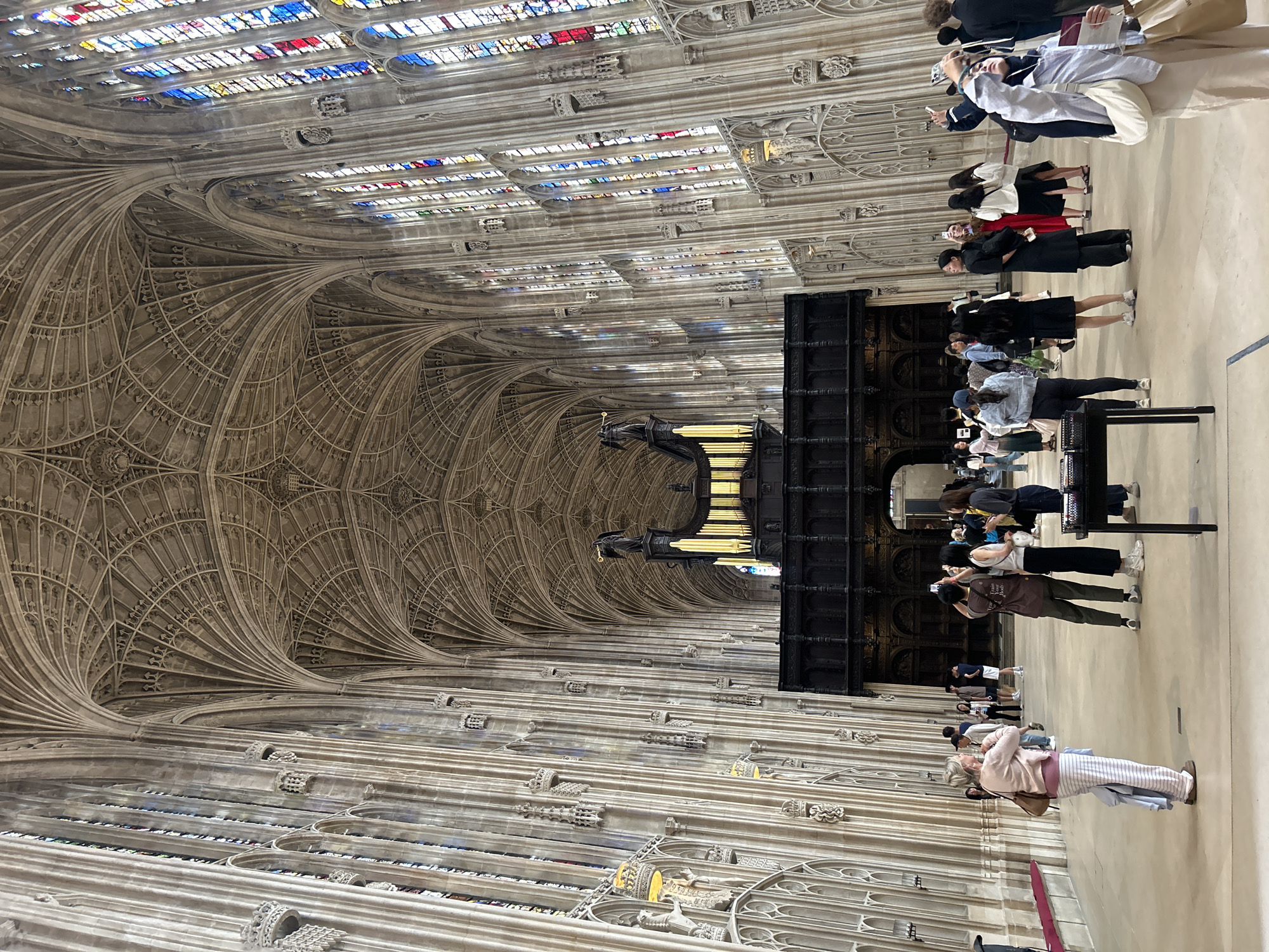 The image depicts the interior of King's College Chapel in Cambridge, United Kingdom. The chapel is characterized by its soaring fan vault ceiling, elaborate stonework, and large stained-glass windows. A pipe organ is visible at the far end of the chapel, above a dark wooden screen. Several tourists are present in the space, some standing and observing, while others are taking photographs with their phones. The floor is light-colored, and the walls are lined with tall, Gothic-style windows.