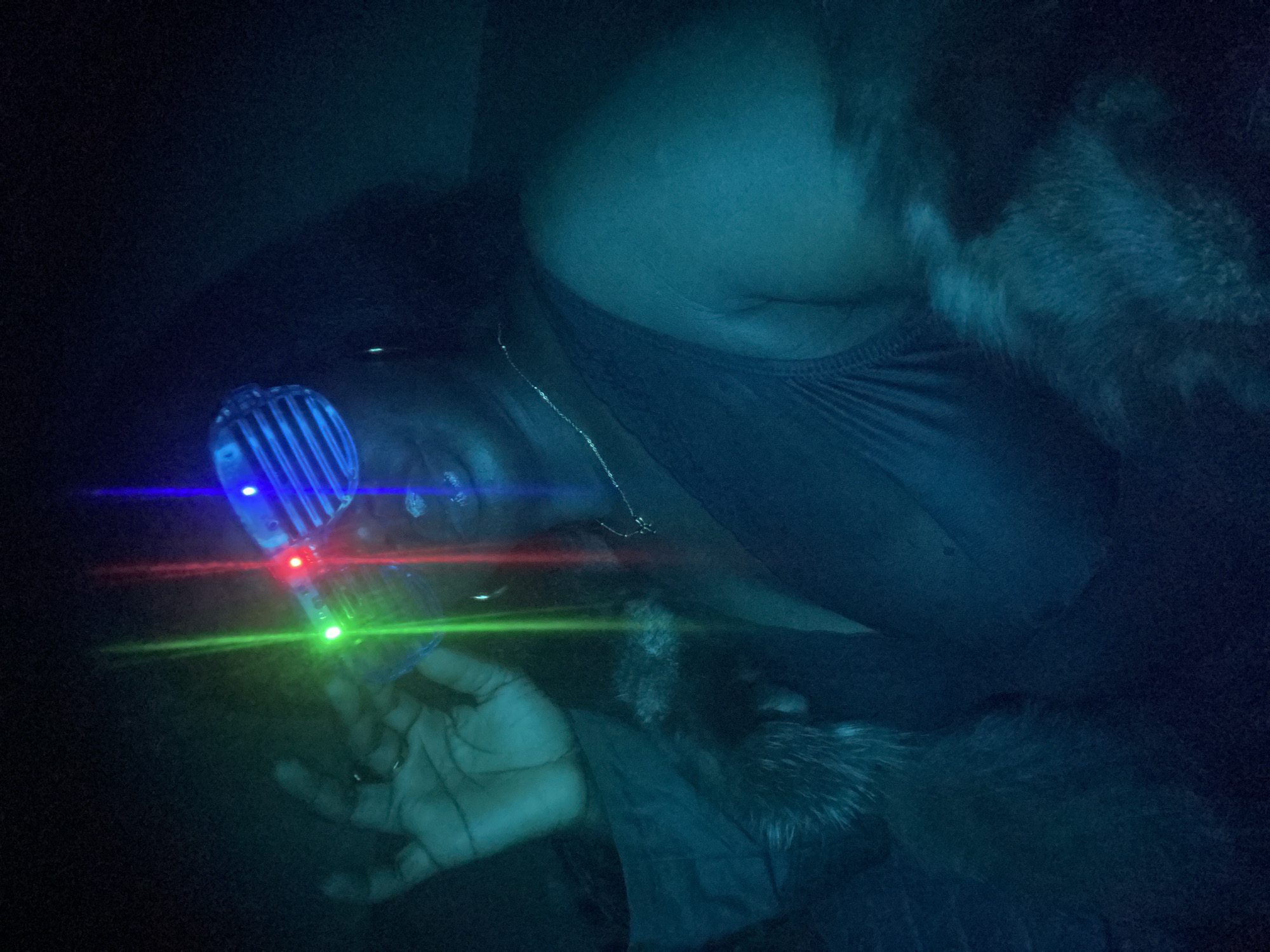 A person is depicted in a dimly lit, dark environment, holding a glowing, handheld device. The individual, whose skin appears dark, wears a dark top and a thin silver necklace. Their face is partially obscured by the blue light emitted from the device, but their left hand, with visible fingers, is clearly holding the object.
The primary object is a futuristic-looking device with a ribbed or textured surface, glowing with bright blue light at its top. From this device, distinct horizontal beams of red and green light emanate, cutting across the dark space. Another blue light source is visible on the device itself.
A dark, furry texture is noticeable near the person's right shoulder and extending towards the lower right of the frame, suggesting a fur-lined garment or perhaps an animal. The overall lighting is very low, creating a mysterious and atmospheric mood, dominated by the artificial colored lights.
The activity involves the person holding and seemingly operating this light-emitting device, which projects colorful lasers into the dark space. The scene appears to take place indoors, likely at night or in a controlled low-light setting, possibly for entertainment or artistic purposes. There are no specific location cues visible within the frame, but the context indicates the location is Maigizo, Nigeria. No text is visible in the image.