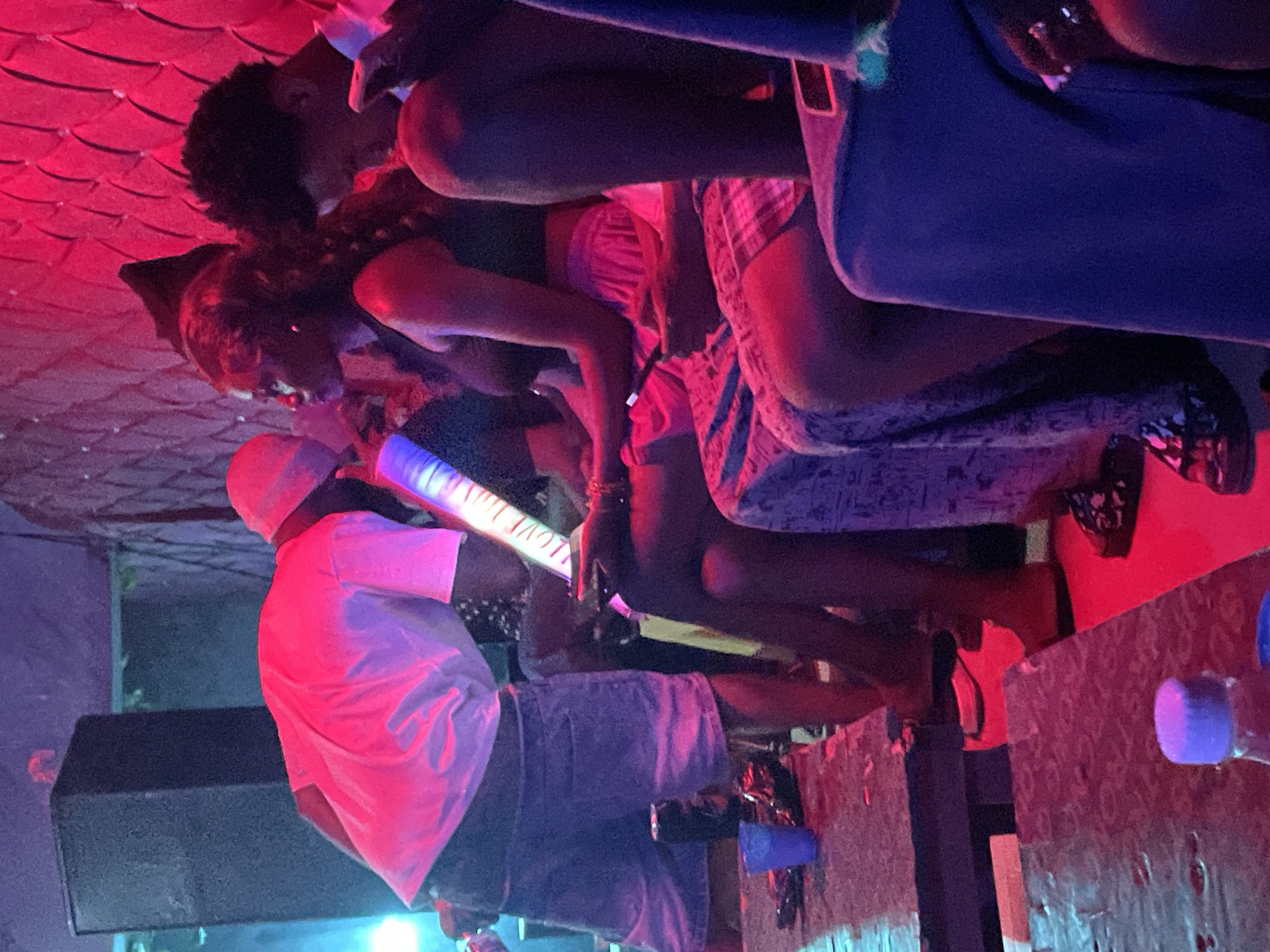 This media file depicts a lively scene in Durumi, Nigeria, likely at night given the artificial lighting. The image is dominated by people and objects in a dimly lit, possibly indoor setting, with red and blue hues suggesting a party or event atmosphere.
The central focus is a group of individuals, some of whom are engaged in close interactions, hinting at a social gathering. One person in the foreground is wearing a white t-shirt and a light-colored hat, facing away from the camera and holding what appears to be a glow stick or light-up toy. Another individual, also seen from behind, is wearing a blue garment. In the background, several other figures are visible, their forms partially obscured by the lighting and framing. There are glimpses of checkered patterns on clothing and a patterned fabric, possibly a seat cover or blanket.
The scene appears to be set in a casual environment, with what looks like a speaker or sound equipment visible on the left side of the image. A table with a textured surface is present on the right, holding a blue cup and possibly other items. The overall mood seems to be one of celebration and enjoyment. There is some visible text on the light-up object being held, which appears to read "LOVE T". The red, textured ceiling above suggests a unique architectural detail or decoration within the venue. The image is oriented in a way that makes the subjects appear to be upside down.
