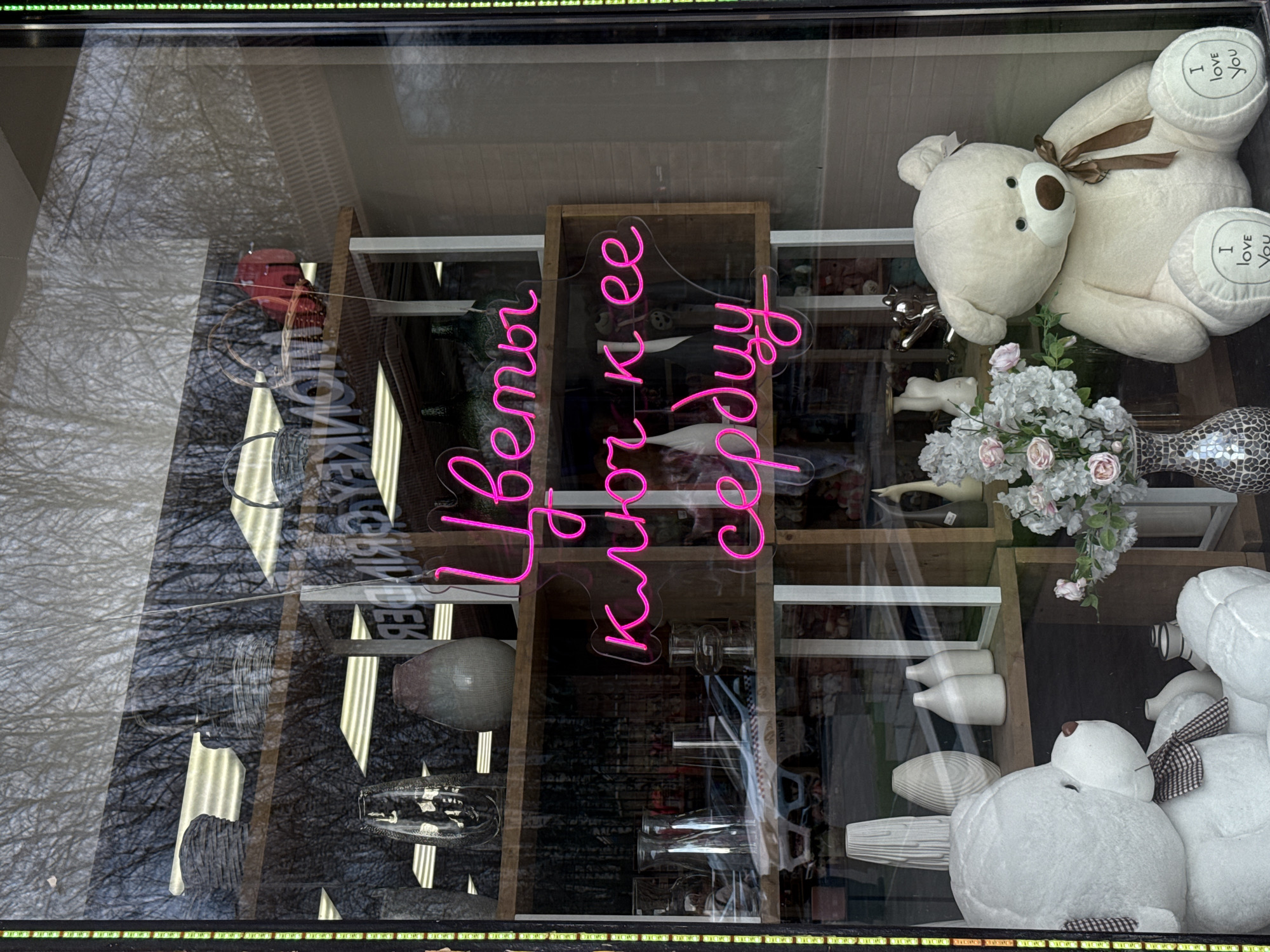 The image shows a storefront display in Perm, Russia, as evidenced by the Cyrillic script in the neon sign. The display includes shelves arranged in a rectangular configuration. On the shelves, various decorative items are visible, including vases, figurines, and plush teddy bears. One of the teddy bears has "I love you" written on the feet. The neon sign, glowing in pink, has the words "Цветы ключ к ее сердцу" meaning Flowers are the key to her heart. There is also a reflected image of trees outside.
