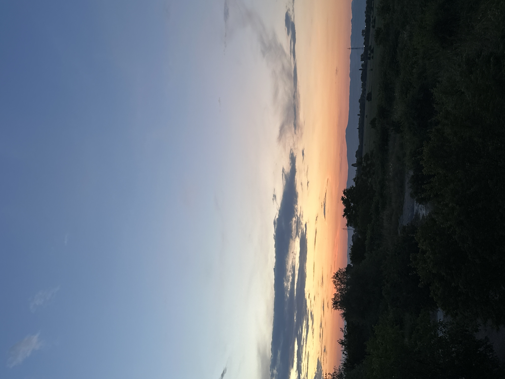 This is a photograph of a sunset over a rural area. The sky is a gradient of colors, from pale blue at the top to orange and pink near the horizon. There are some wispy clouds scattered across the sky. In the foreground, there is a dark silhouette of trees and vegetation. In the distance, there are rolling hills and a few power lines. The overall impression is one of a peaceful evening. The location is the outskirts of Sofia, Bulgaria.