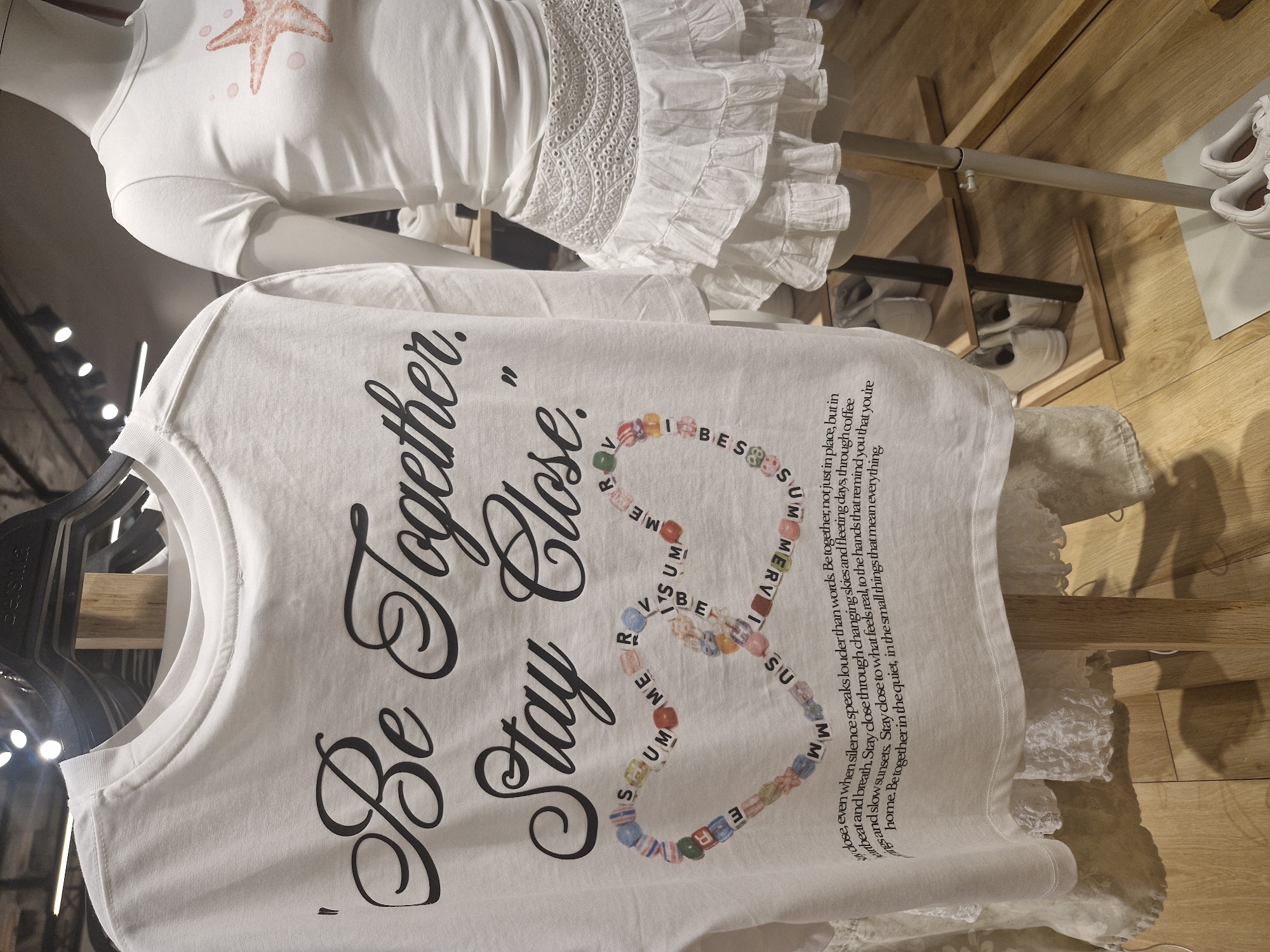 The image captures a close-up view of two white garments displayed in an indoor setting, likely a retail store or a wardrobe.
In the foreground, a white short-sleeved t-shirt hangs, featuring prominent black script text that reads, "Be Together. Stay Close." Below this text, a heart-shaped outline is formed by colorful, cube-shaped beads, which spell out "SUMMER VIBES." To the right of the heart, a paragraph of smaller black text is printed on the t-shirt, stating: "close, even when silence speaks louder than words. Be together, not just in place, but in heart and breath. Stay close through changing skies and fleeting days, through coffee mornings and slow sunsets. Stay close to what feels real, to the hands that remind you that you're home. Be together in the quiet, in the small things that mean everything."
In the background, on a white mannequin, another white top is displayed. This top has a light orange starfish graphic on its chest. The lower part of the mannequin's garment is adorned with white eyelet lace and ruffles.
The setting includes a wooden floor and shelves, where pairs of white sneakers are visible. A grey metal clothing rail extends horizontally across the middle ground. Bright, circular lights can be seen in the upper left of the frame, suggesting overhead lighting. The overall color palette is dominated by white, black, and natural wood tones, accented by the colorful beads on the t-shirt. The text on the foreground t-shirt conveys themes of intimacy, connection, and appreciation for small, meaningful moments.