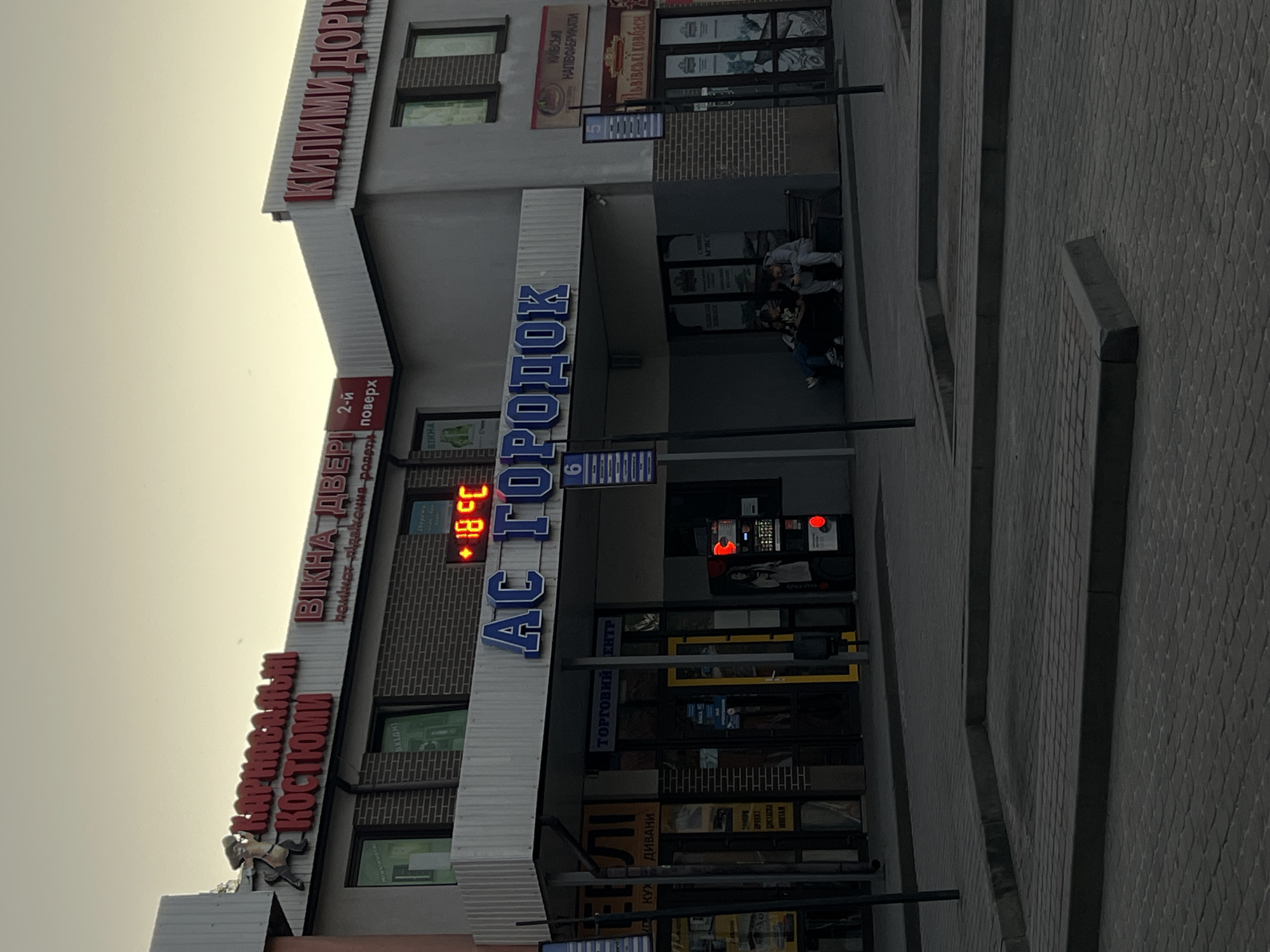 The image shows the exterior of a multi-story commercial building in Horodok, Ukraine. The building has multiple businesses including Килими Дорі (carpets), a costume shop, and Vikna Dveri (windows and doors) located on the second floor. A sign reading "AC Городок" is prominently displayed. A digital temperature display reads +18 degrees Celsius. People are seen sitting in the entryway of the building. The street and sidewalk are paved with interlocking pavers. There are streetlights along the side walk.