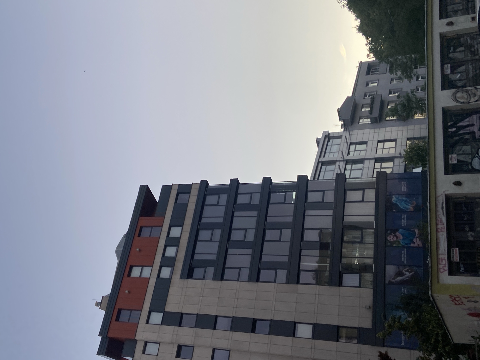 The image shows a low-angle view of several modern buildings against a pale, overcast sky. A small airplane is visible in the distance. The buildings are a mix of glass, concrete, and reddish-brown panels. One building on the left has a section with orange and beige cladding and many dark-framed windows. To its right, another building is predominantly grey with a large facade featuring advertisements. These advertisements include images of people engaged in activities like swimming and smiling, with accompanying text such as "LIFESTYLE MEDICINE," "MEDICAL IMAGING," and "CARE." Further to the right, a building with a graffiti-covered exterior is visible. Trees are interspersed between the structures, and the overall impression is an urban environment. The lighting suggests it is daytime, possibly late afternoon or early morning, with diffused sunlight. The sky is a uniform light grey, indicating cloudy weather. The presence of Polish text on the advertisements and the provided location context confirm the setting as Warsaw, Poland.