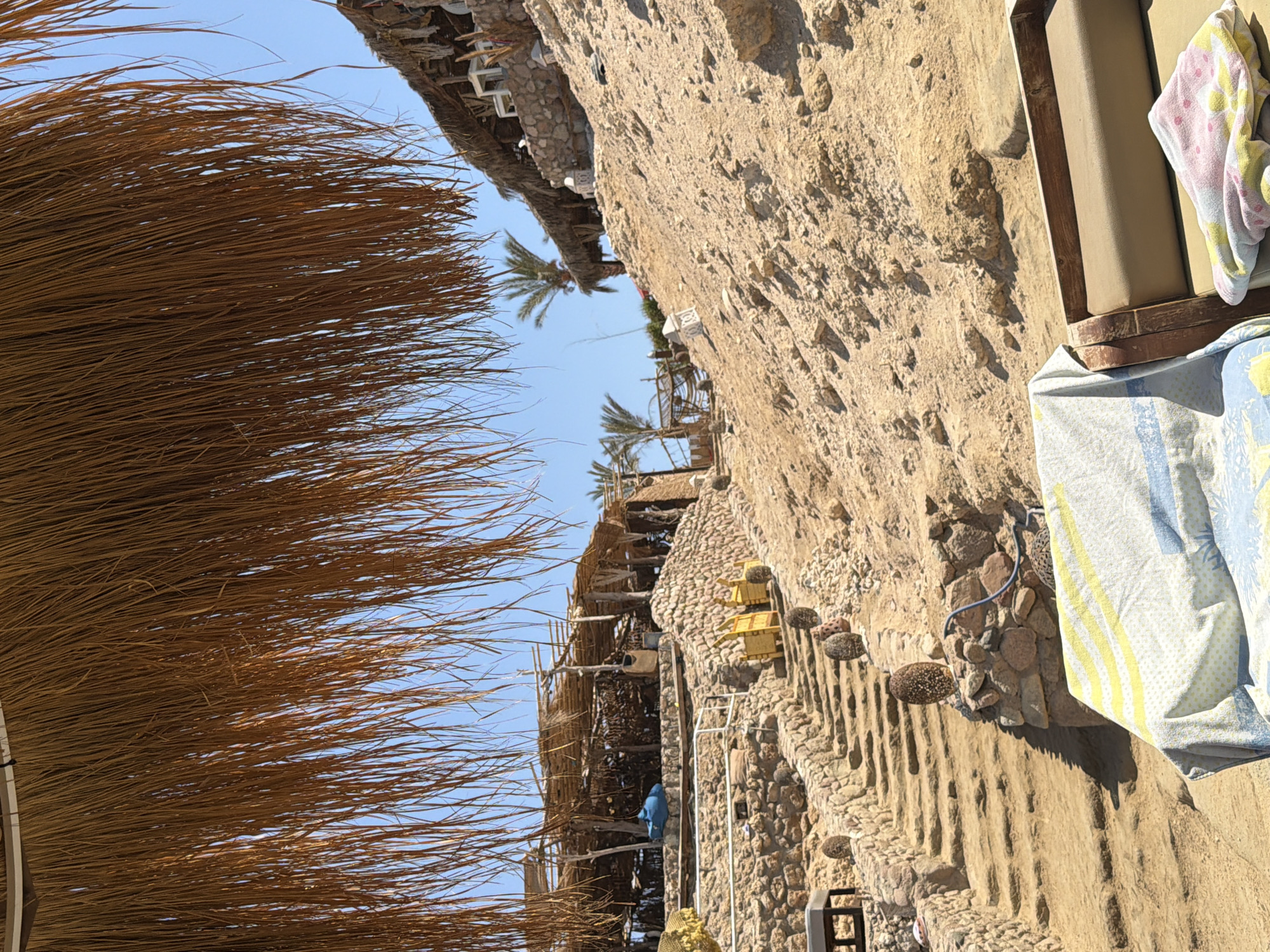 The image depicts a sun-drenched outdoor scene on a sandy hillside, likely a beach resort area in Sharm El Sheikh, Egypt. A thatched roof overhang, made of dried palm fronds, dominates the foreground, casting a shadow. Below, a stone staircase winds its way up the rocky terrain. Scattered along the stairs are decorative, egg-shaped objects. At the top of the hill, rustic structures with thatched roofs and open sides are visible, resembling a restaurant or seating area. Palm trees are present in the background, suggesting a warm, coastal environment. In the immediate foreground, a lounge chair is partially covered with towels, indicating a recreational space. The sky is clear and blue. The overall impression is of a tranquil, resort-like setting by the sea.