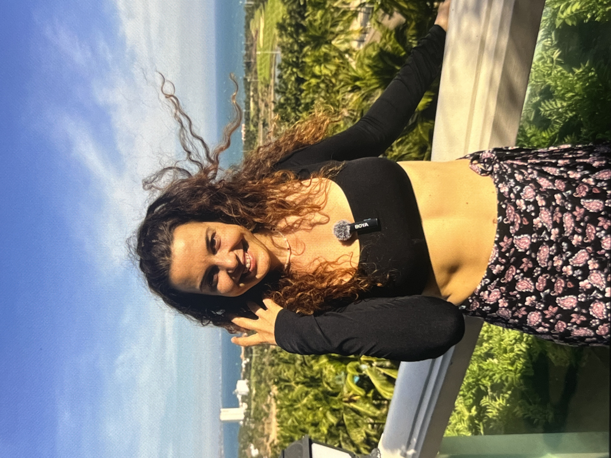 A woman with curly brown hair stands on a balcony overlooking a tropical landscape. She is wearing a black long-sleeved crop top and a black skirt with a pink floral pattern. She is smiling and has a microphone clipped to her top. The background consists of palm trees, a golf course, buildings, and the ocean under a clear blue sky. The scene is lit by sunlight.