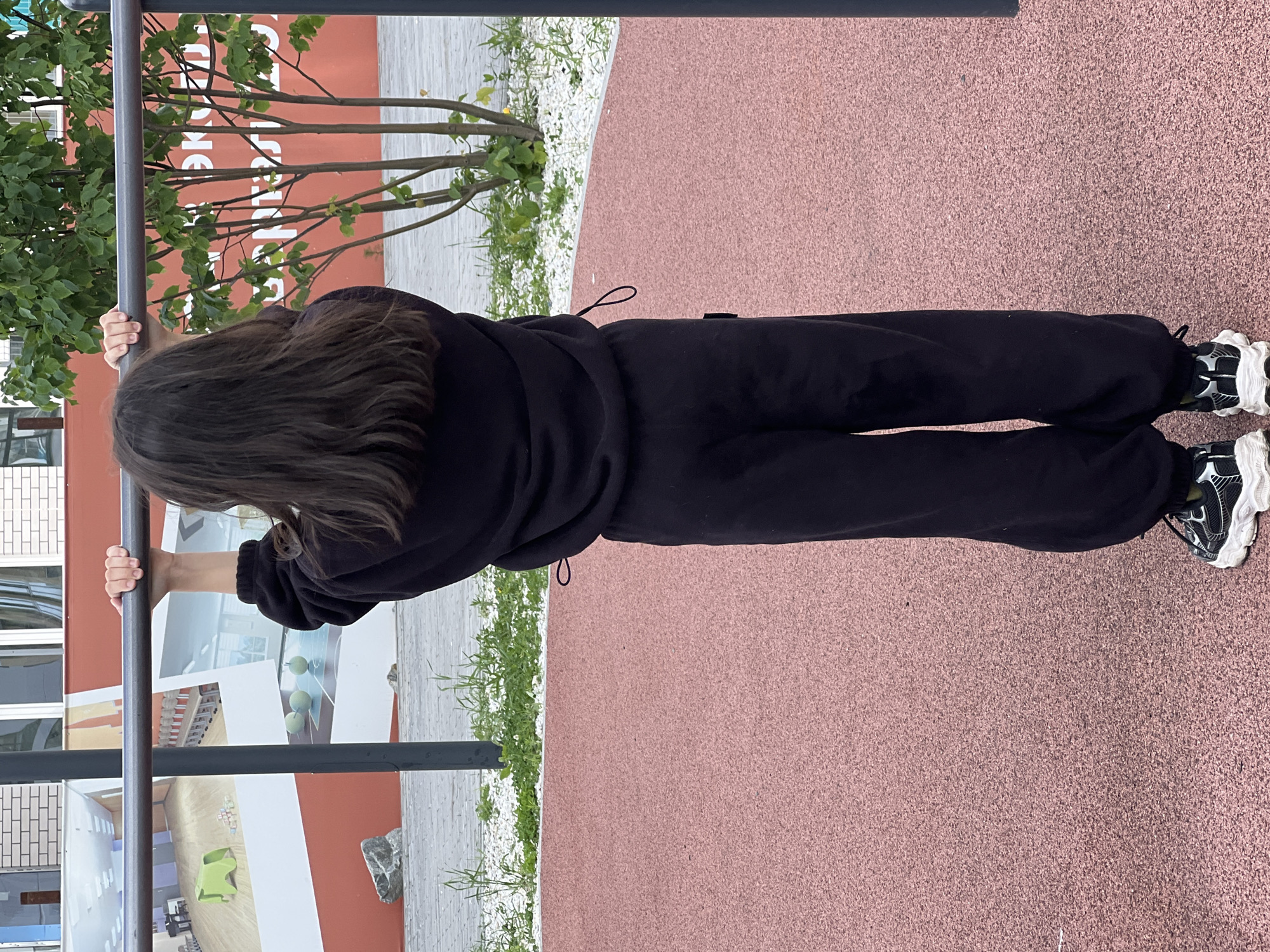 An unidentified child, seen from behind, stands on a reddish-pink rubberized athletic surface, holding a horizontal dark grey metal bar. The child has long, dark brown hair and is dressed in a black long-sleeved top with a visible drawstring loop on the left side, and black loose-fitting sweatpants. Black and white athletic shoes with prominent white soles are worn on both feet.
In the background, behind the bar, a building features a red-painted lower section and white brickwork above. Large banners or murals are affixed to the red wall. The left banner depicts an interior room with a green animal-shaped chair and a piano. The right banner shows what appears to be a classroom or assembly hall with rows of seating and a whiteboard, alongside three light green spheres on a surface. Between and around these banners, Russian text is visible on the red wall, with the word "ЕКАТЕРБУРГ" (Yekaterinburg) clearly identifiable, indicating the location in Yekaterinburg, Russia.
A young deciduous tree with multiple slender trunks and green leaves stands to the right of the child, partially obscuring the background wall. Beyond the rubberized surface, a grey paved area, interspersed with white gravel and green weeds, is visible. Grey vertical poles support the horizontal bar. The scene is outdoors under an overcast sky.