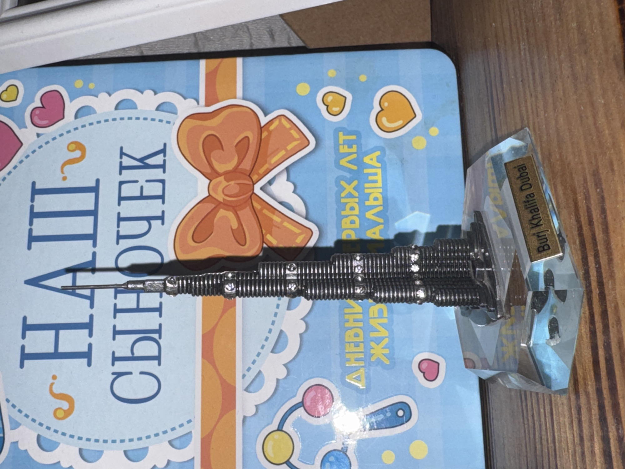 The image shows a miniature model of the Burj Khalifa tower placed on a wooden surface. The model is dark gray with silver accents and is mounted on a clear, faceted base. A small brass plaque on the base reads "Burj Khalifa Dubai." Behind the model, a blue card with decorative elements is visible. The card features the text "НАШ СЫНОЧЕК" in large blue letters, along with images of hearts, a bow, and a baby rattle. The overall scene appears to be a personal keepsake or decoration. The location, Яромінскі сельскі Савет, Belarus, is a rural administrative division and does not directly relate to the objects depicted in the image itself.