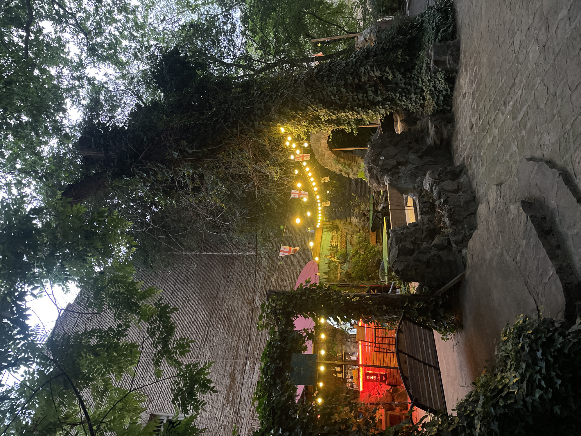 The image depicts an outdoor setting at dusk, likely in the city of Tbilisi, Georgia, based on the Georgian flags strung between lights. The scene includes a building with a textured wall partially obscured by foliage, with trees overhead forming a dense canopy. Strings of lights with Georgian flags are draped across the scene, leading towards a more brightly lit area that seems to be a bar or outdoor seating area, noticeable by the red lighting. Stone structures are also present, possibly part of a retaining wall or decorative feature. The ground surface appears to be a mixture of stone and concrete. The overall atmosphere suggests a relaxed, evening setting in an urban environment with a natural, overgrown aesthetic.