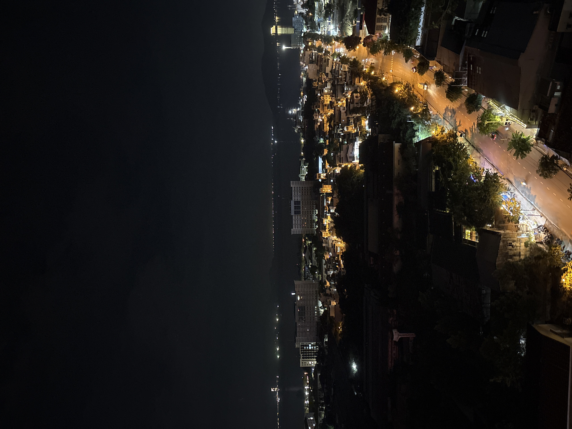 This is a nighttime aerial view of a city, likely Nha Trang, Vietnam, looking out towards a body of water. The foreground is dominated by dark buildings and trees, with some illuminated windows visible. A paved road runs through the mid-ground, with streetlights illuminating the asphalt and casting long shadows. Several vehicles, including cars and motorcycles, are on the road. Further into the scene, there are more buildings of various sizes, some appearing to be hotels or apartment complexes, all lit up. Beyond the city lights, a wide expanse of dark water stretches to the horizon, dotted with numerous small lights that could indicate boats or structures on the water. In the far distance, hills or mountains are silhouetted against the dark sky. The overall impression is a vibrant, illuminated cityscape at night against a dark natural backdrop.